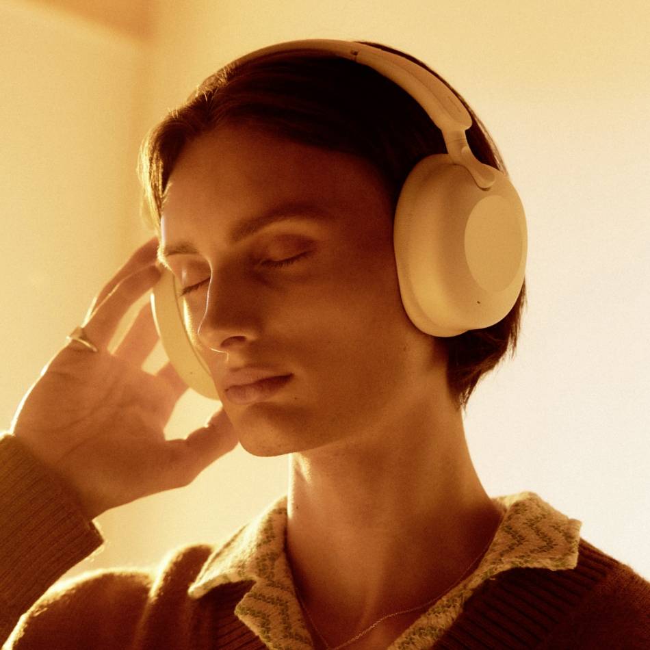 Persoon met witte koptelefoon die muziek geniet en met gesloten ogen ontspant, draagt een trui met kraag. Warm licht.