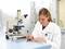 Vrouw in laboratorium schrijft op een objectglaasje naast een microscoop, draagt een laboratoriumjas. Achtergrond toont laboratoriumapparatuur.