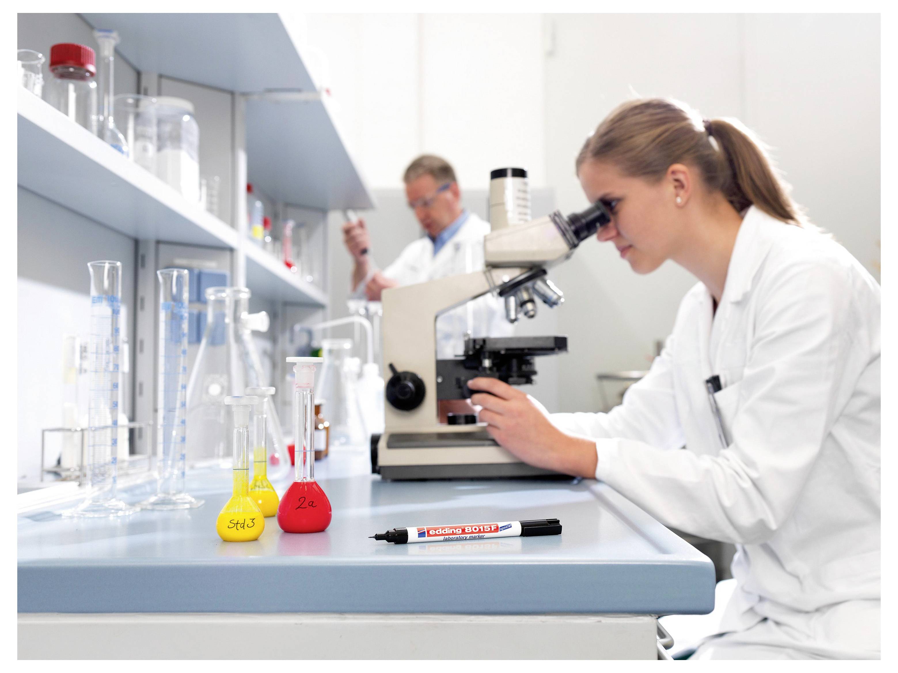 Wetenschappers in een laboratorium, met een vrouw die een microscoop gebruikt aan een werkbank omringd door flessen, bekers en chemicaliën.