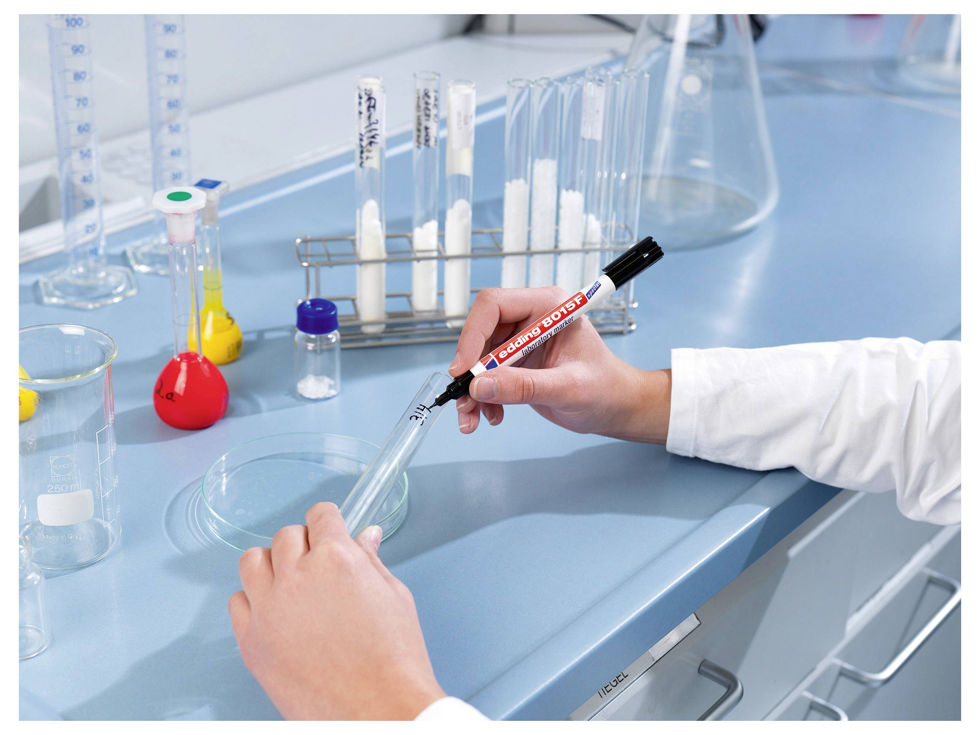 Een persoon in een wit laboratoriumjas die een duidelijke reageerbuisje markeert met een marker op een laboratoriumbank, met kolf, bekers en monsterbuisjes op de achtergrond.