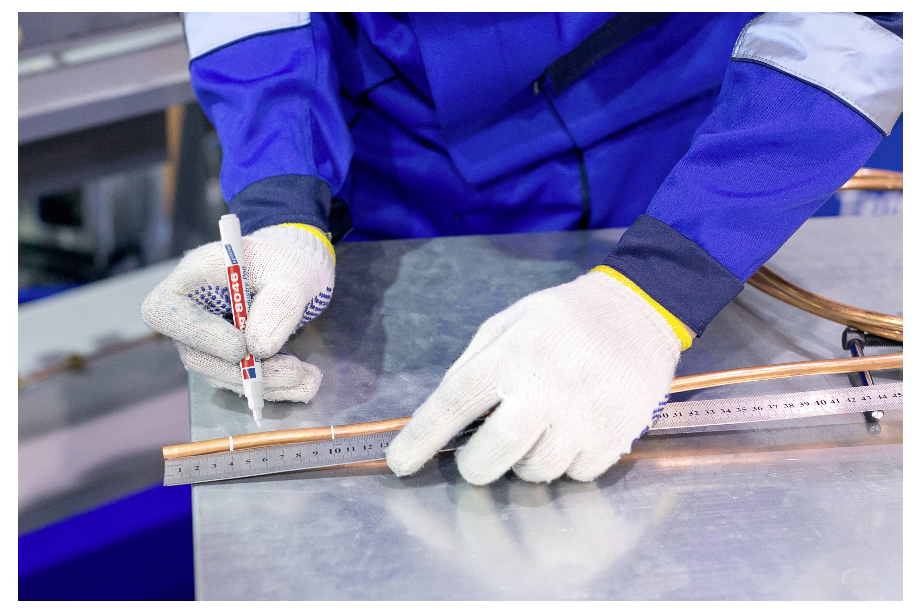 Werknemer in blauwe werkoverall en handschoenen markeert een koperen buis naast een metalen liniaal op een werkbank.