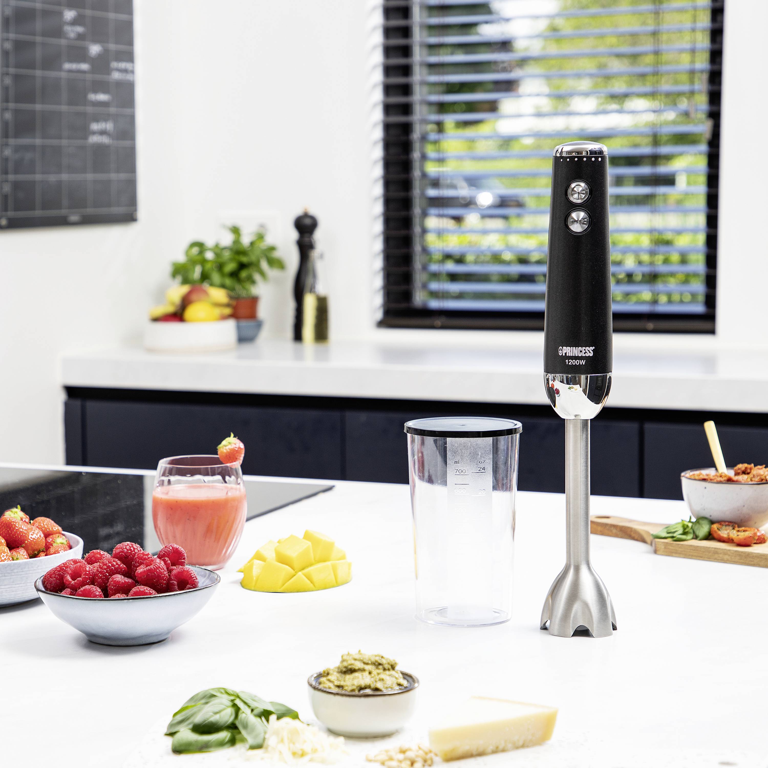 Een staafmixer staat op een aanrecht, omringd door verse ingrediënten zoals aardbeien, mango en Parmezaanse kaas; klaar voor bereiding.