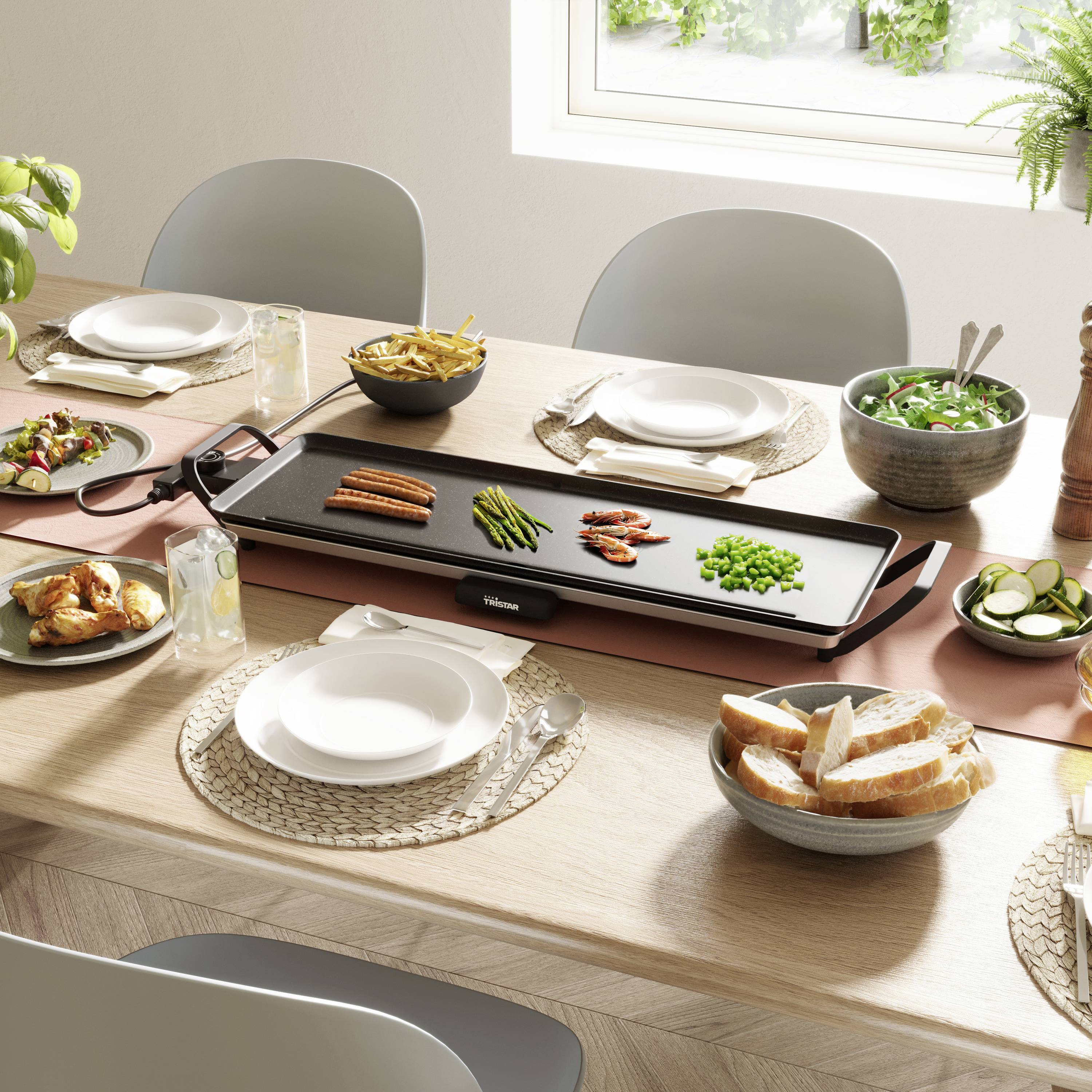 Een gedekte tafel met een elektrische grill in het midden, groenten en vlees erop, omringd door servies en brood.