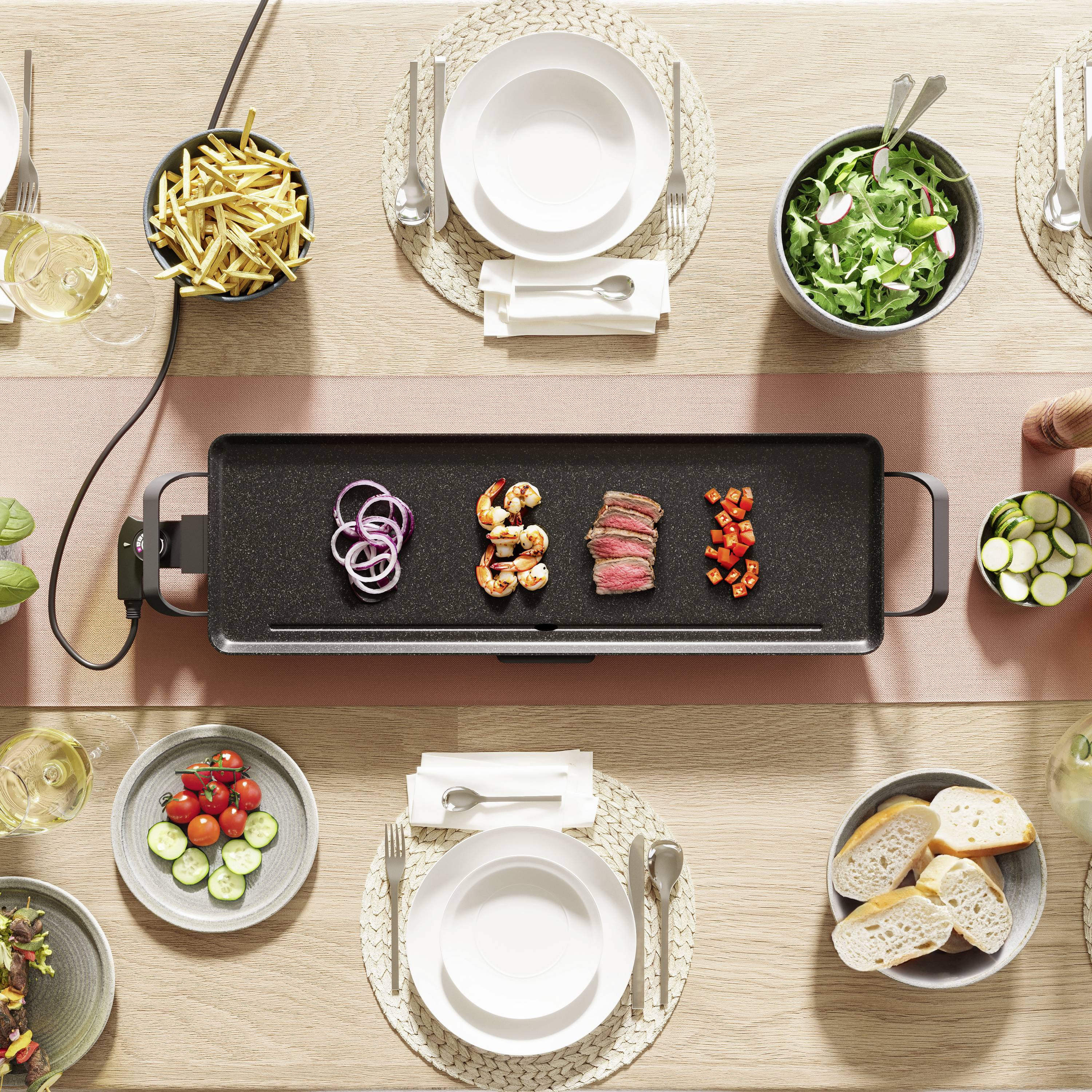 Een tafel met een grillplaat, daarop garnalen, vlees en groenten. Eromheen salade, friet, brood en lege borden met bestek.