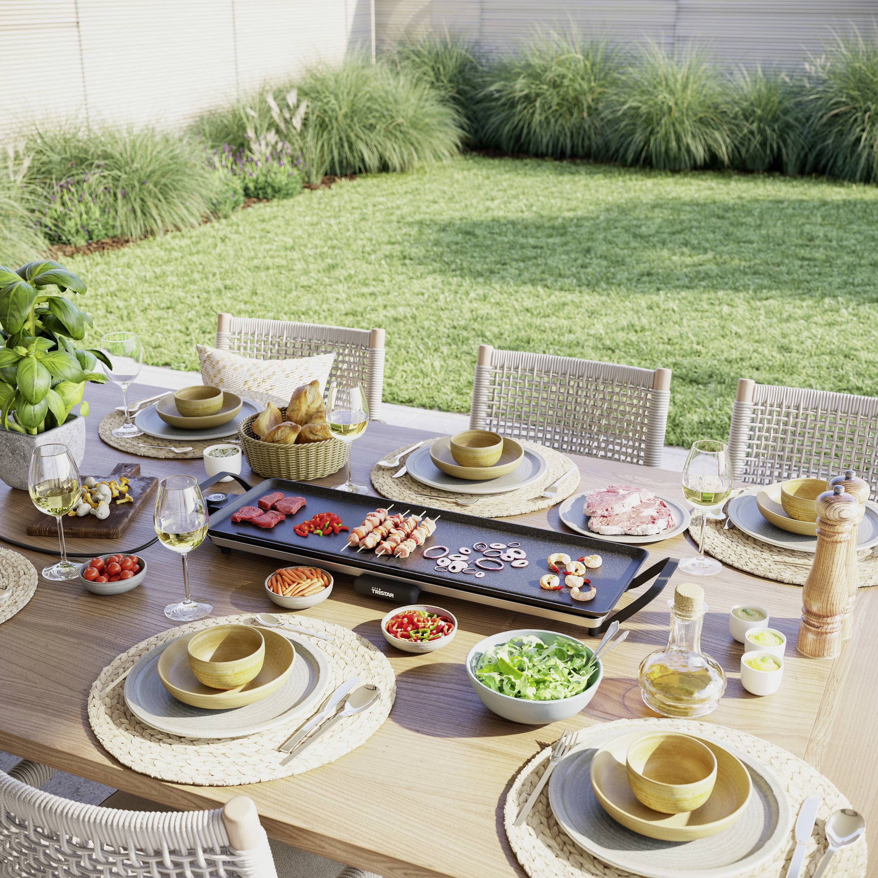 Een gedekte eettafel in de tuin met een grillplaat, verschillende ingrediënten, servies en dranken. Op de achtergrond gras en struiken.