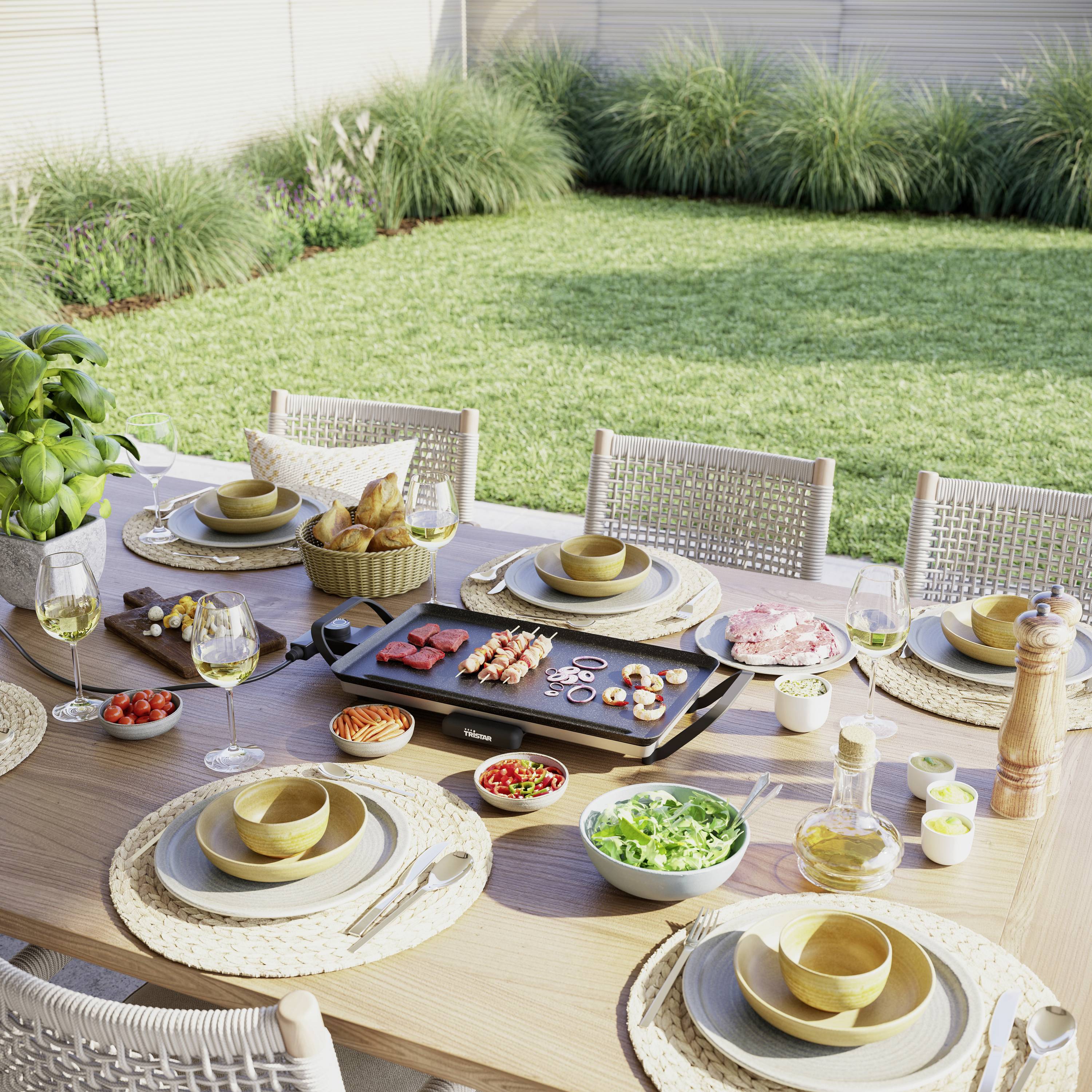 Een gedekte tuintafel met een grillplaat waarop vlees en groenten worden bereid, omringd door servies en glazen. Op de achtergrond een gazon.