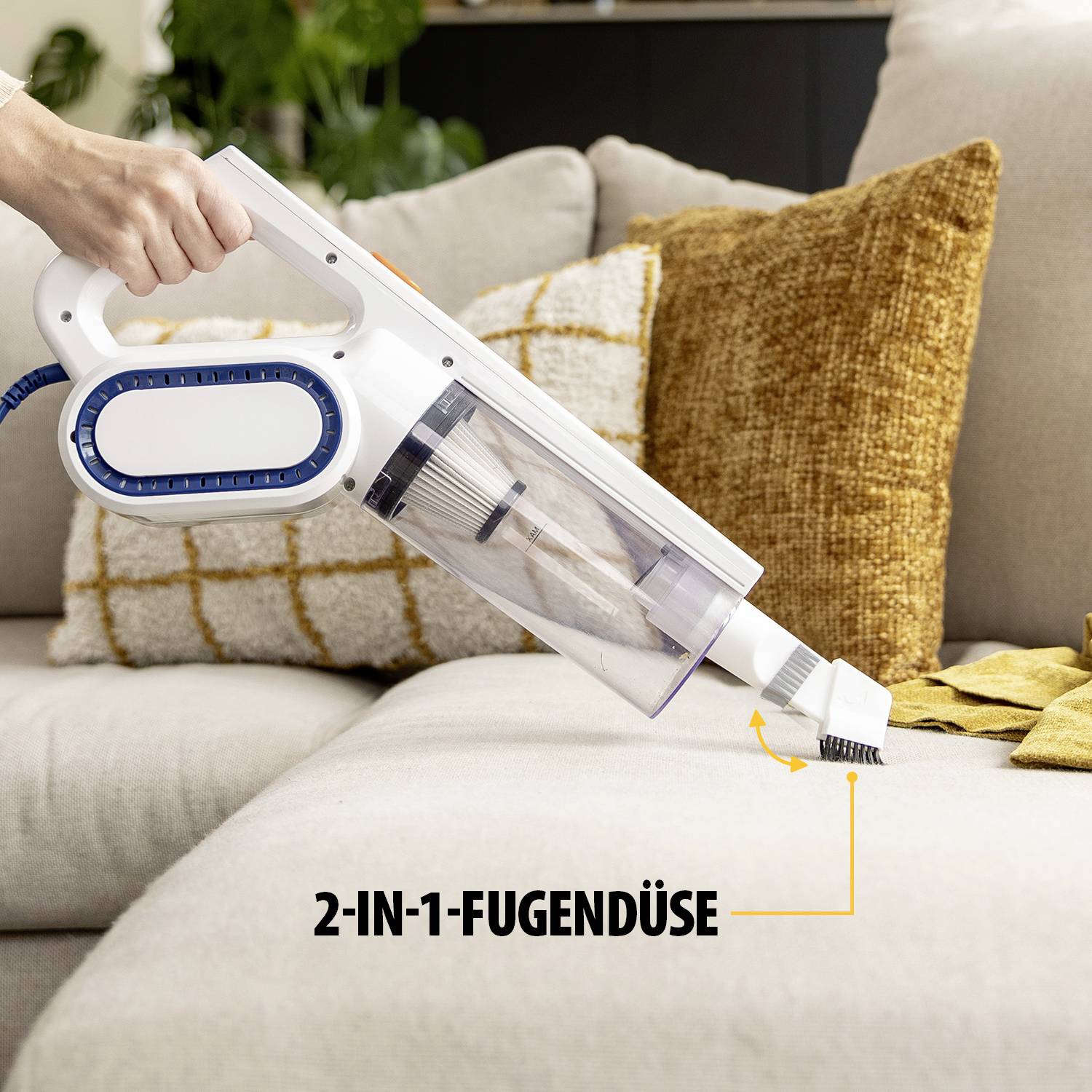 Een persoon gebruikt een draagbare stofzuiger met een speciale 2-in-1 spleetmondstuk om een bank schoon te maken. Op de achtergrond zijn kussens zichtbaar.
