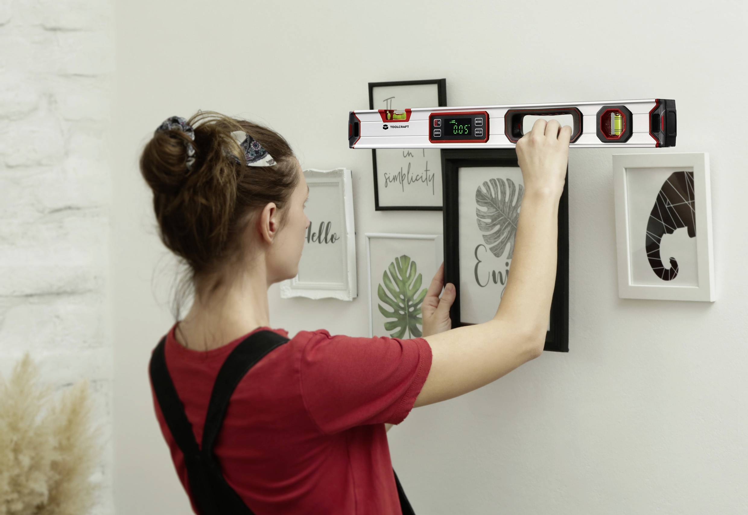 Een persoon hangt schilderijen aan een witte muur op, waarbij hij een waterpas gebruikt voor nauwkeurige uitlijning. Op de achtergrond plantenmotieven.
