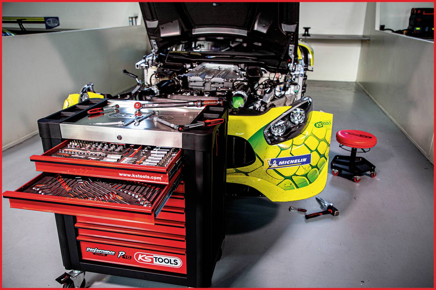 Racewagen met geopende motorkap in werkplaats, geflankeerd door een gereedschapswagen. Mechanische stoel staat ernaast.