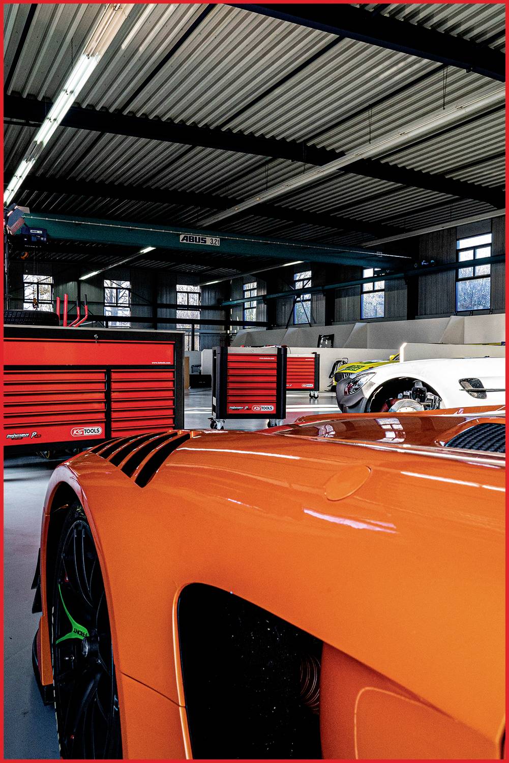 Een oranje auto staat in een werkplaats voor een witte auto en verschillende gereedschapskasten.