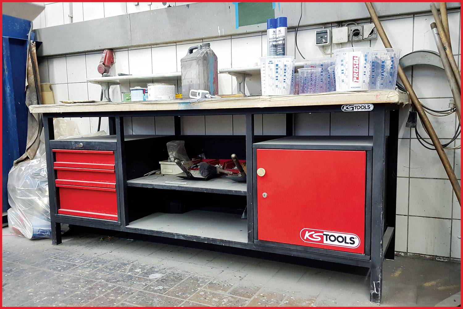 Werkbank met rode lades en een kast, gevuld met gereedschappen en materialen. Hij staat in een werkplaats voor een betegelde muur.