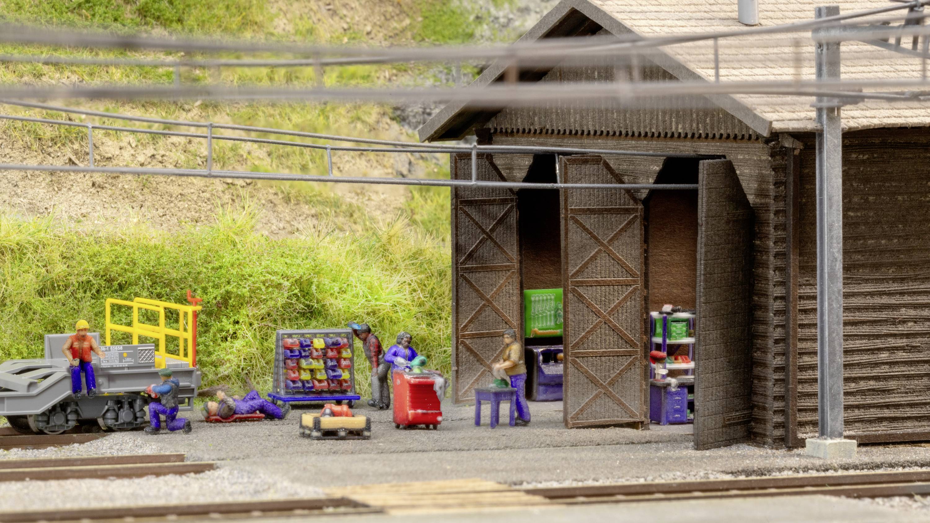 Een miniatuur motoronderhoudsgebied met kleine figuren die werken aan een modelspoorbedrijfsgebouw. Kabels lopen erbovenop.