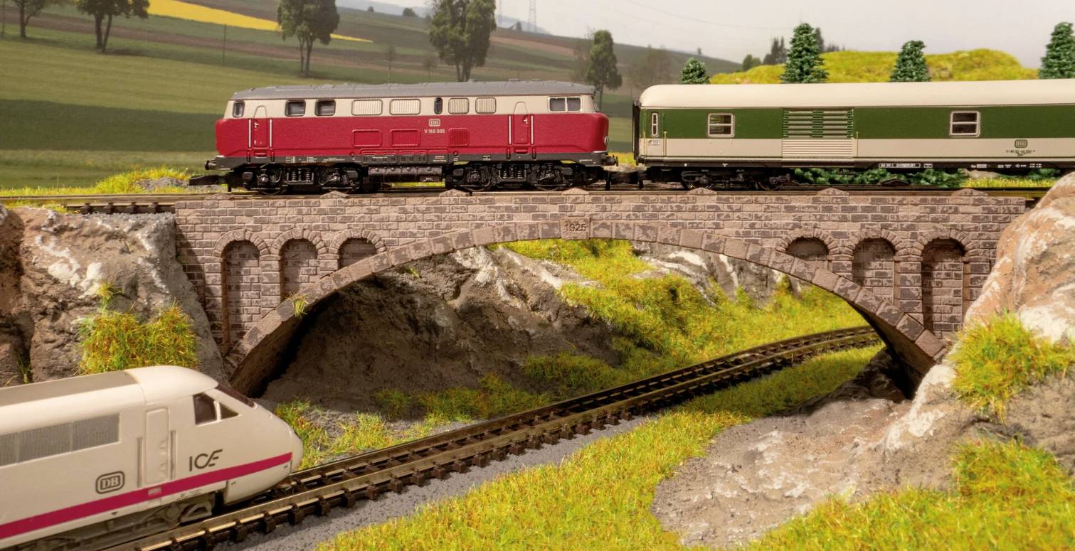 Treinmodel-scenerie met een rode en groene trein over een stenen brug, waarbij daaronder een witte ICE-trein door een schilderachtig landschap rijdt.