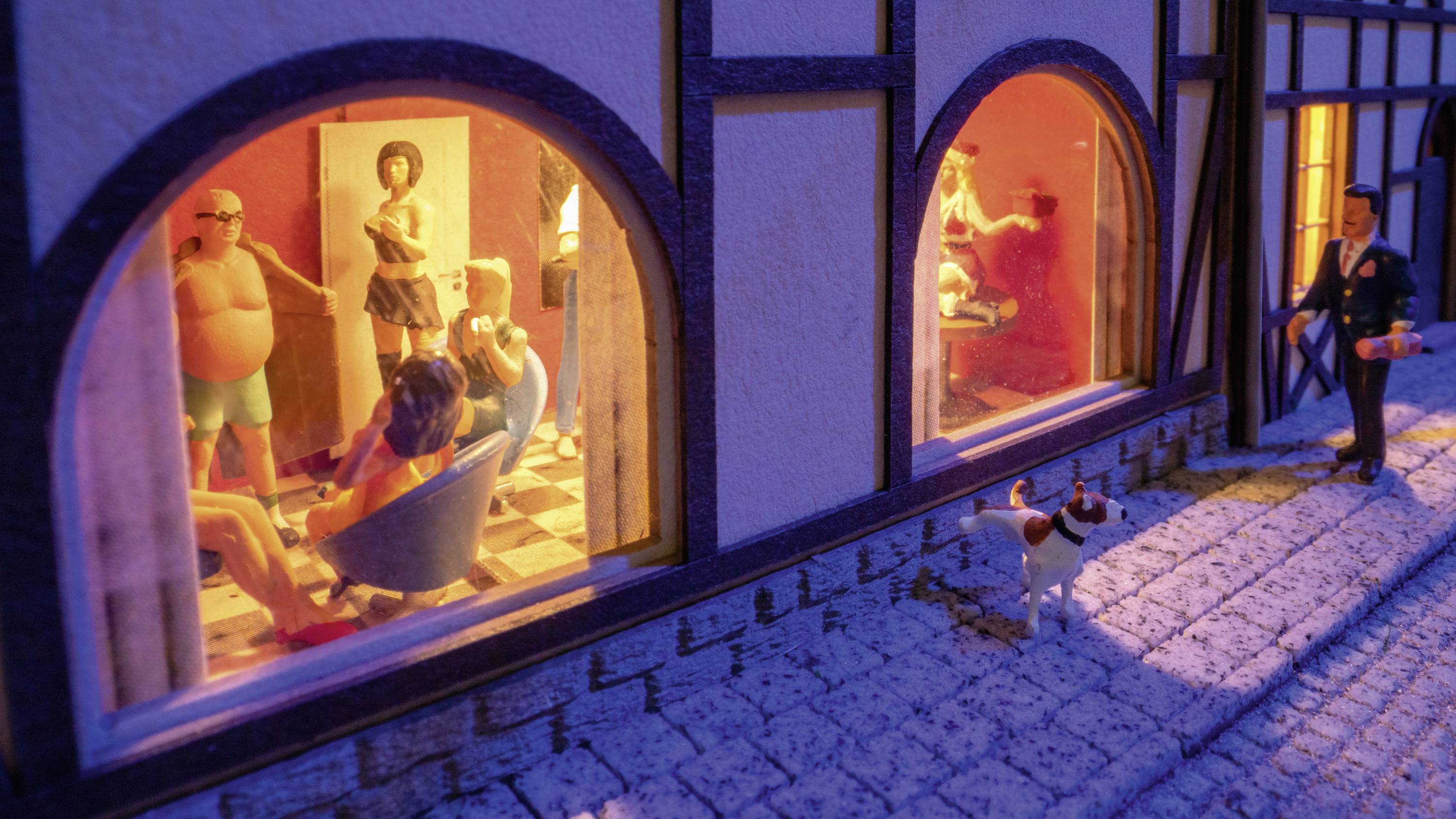 Een straatscène bij nacht toont mensen in verlichte ramen van een huis. Buiten wandelt iemand met een hond.