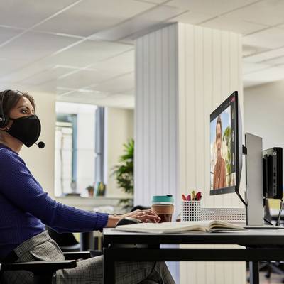 Een persoon met koptelefoon zit aan een bureau en houdt een online vergadering op een computerscherm. Er staan planten op de achtergrond.