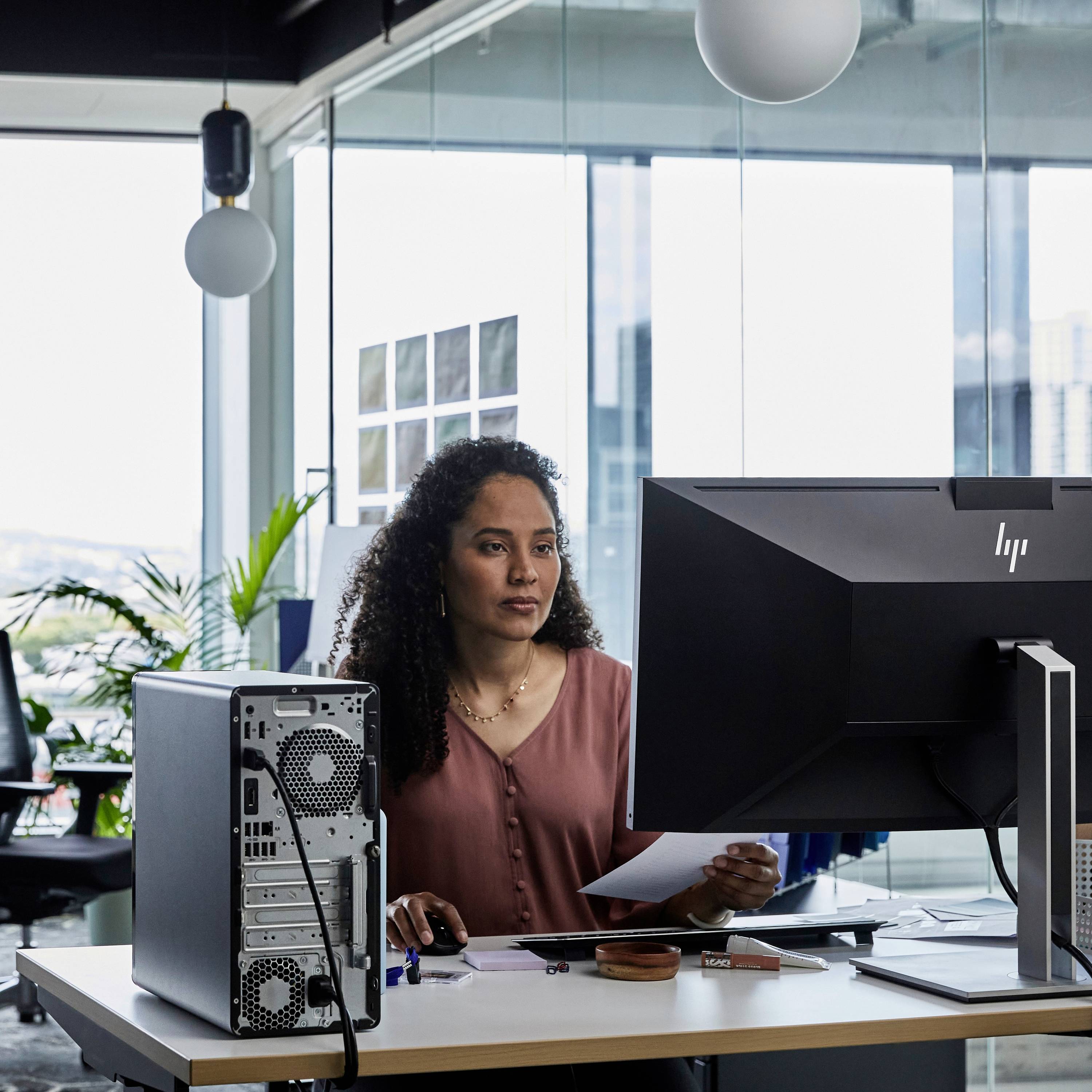 Een vrouw zit aan een bureau met computers in een modern kantoor. Ze kijkt naar een scherm en houdt een pen en papier vast.