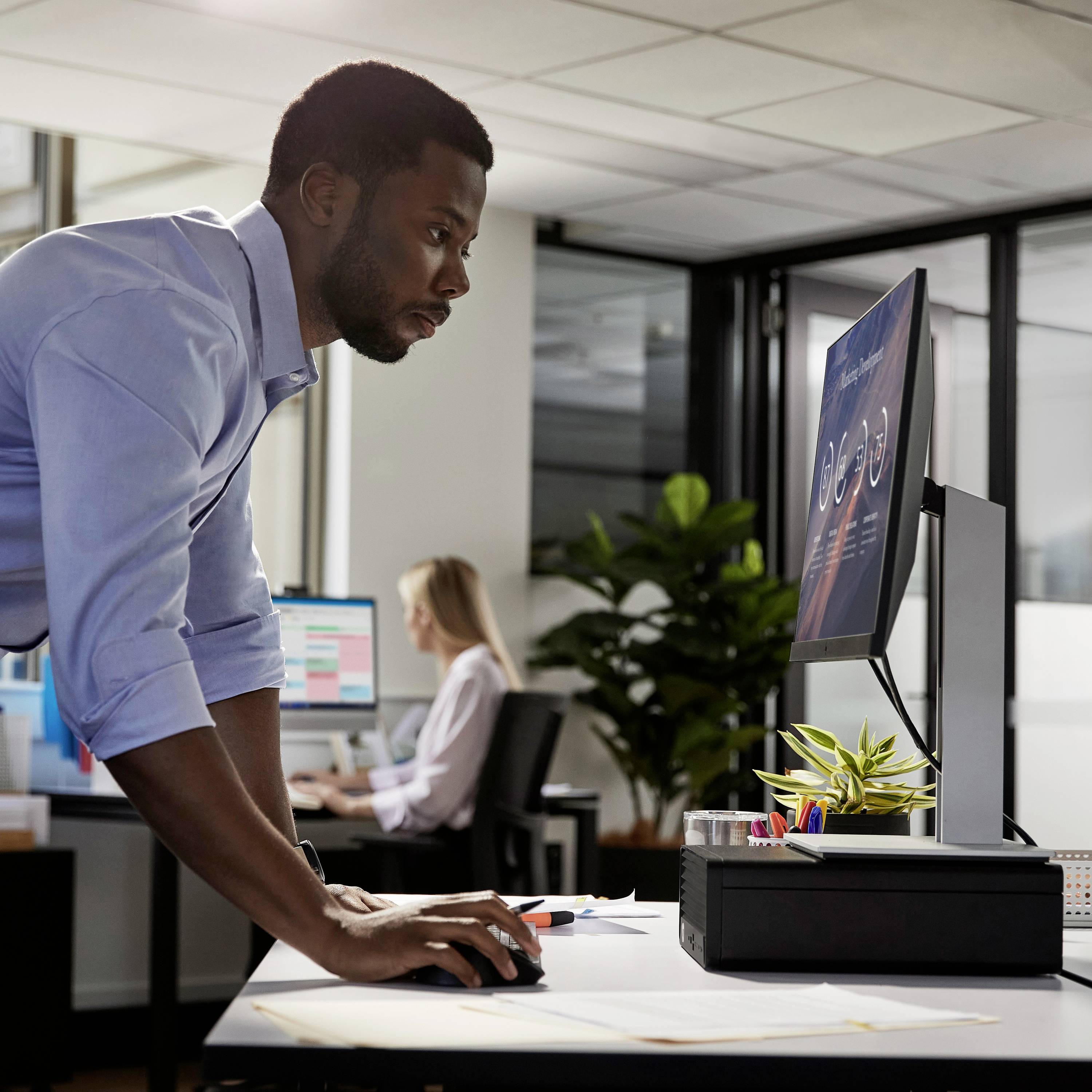 Een man staat bij een bureau in een modern kantoor en kijkt naar een computerscherm. Op de achtergrond werken andere personen.