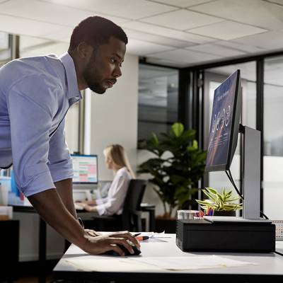 Een man staat bij een bureau in een modern kantoor en kijkt naar een computerscherm. Op de achtergrond werken andere personen.
