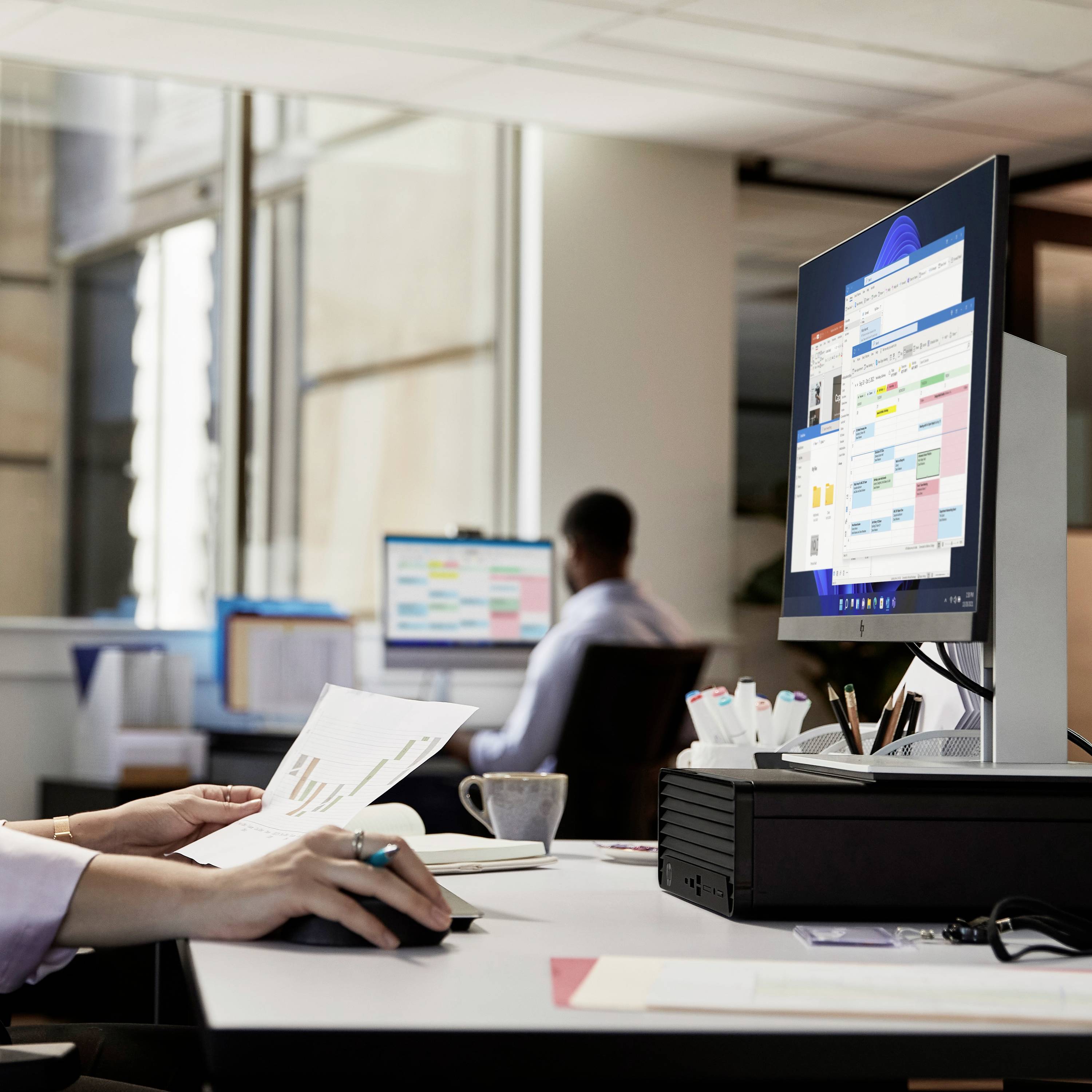 Een kantoor met twee personen aan bureaus die op computers werken. Op het scherm zijn kleurrijke kalenders en planners zichtbaar.