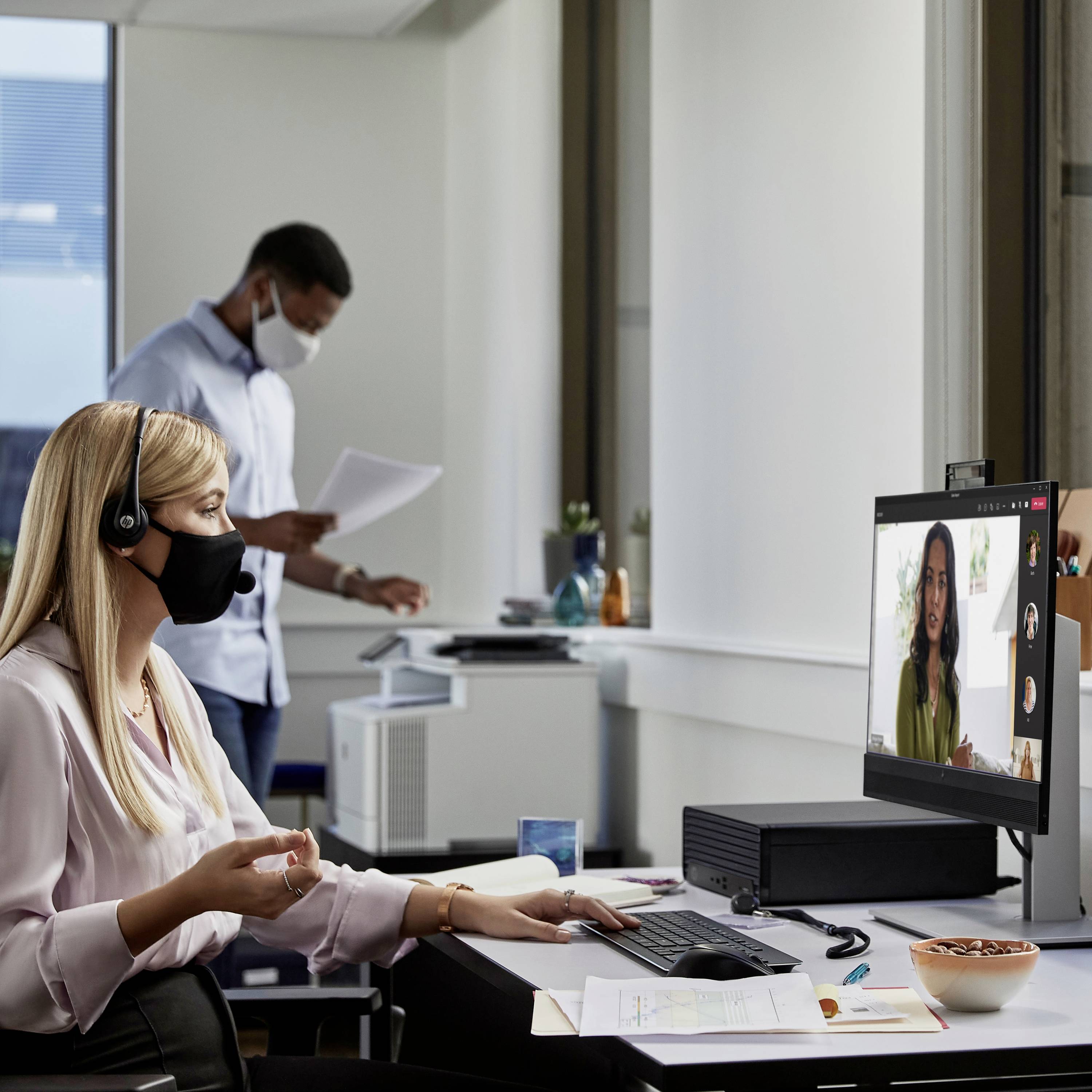 Twee personen op kantoor, een vrouw in een videoconferentie achter de computer, een man met documenten op de achtergrond, beiden dragen mondmaskers.
