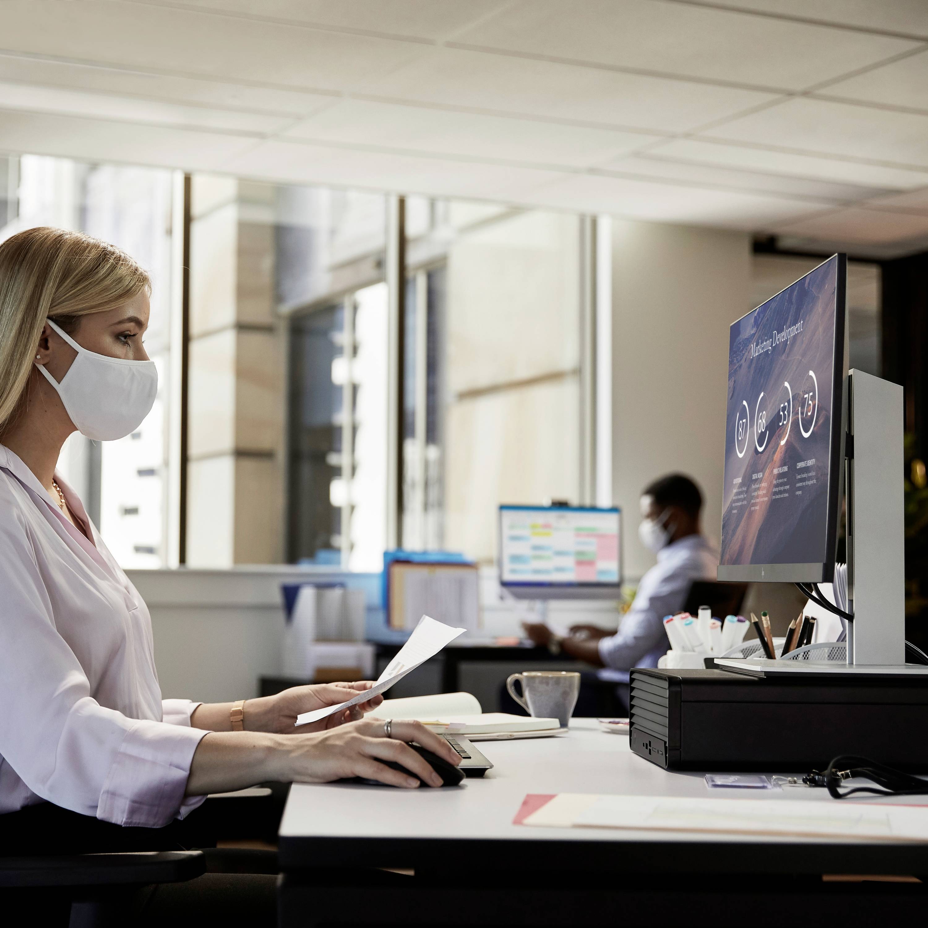 Een vrouw met een masker werkt aan een bureau voor een computerscherm met grafieken. Op de achtergrond bevindt zich nog een medewerker.