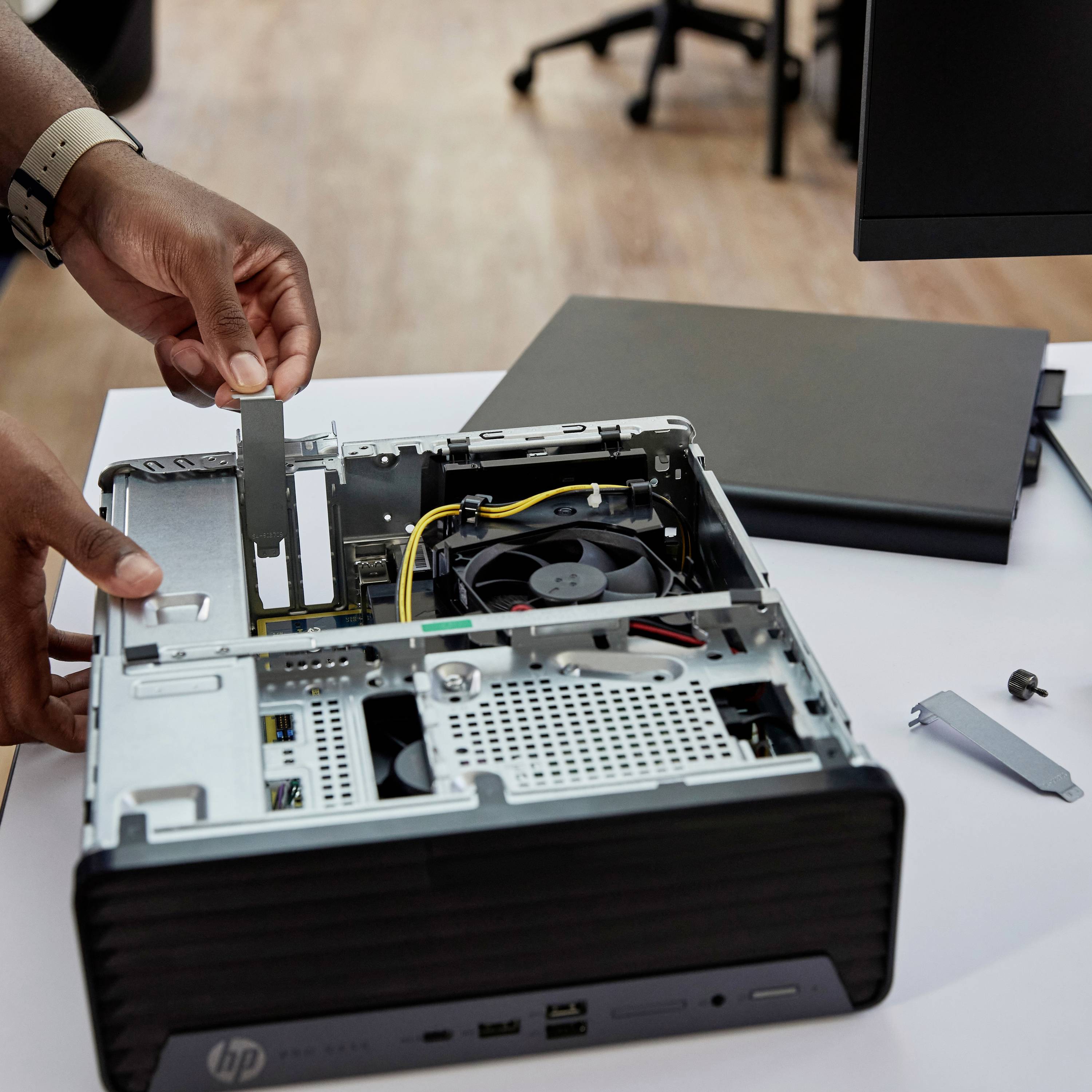 Een persoon installeert een nieuwe hardware-component in een open desktop-computer, met een monitor op de achtergrond op een bureau.