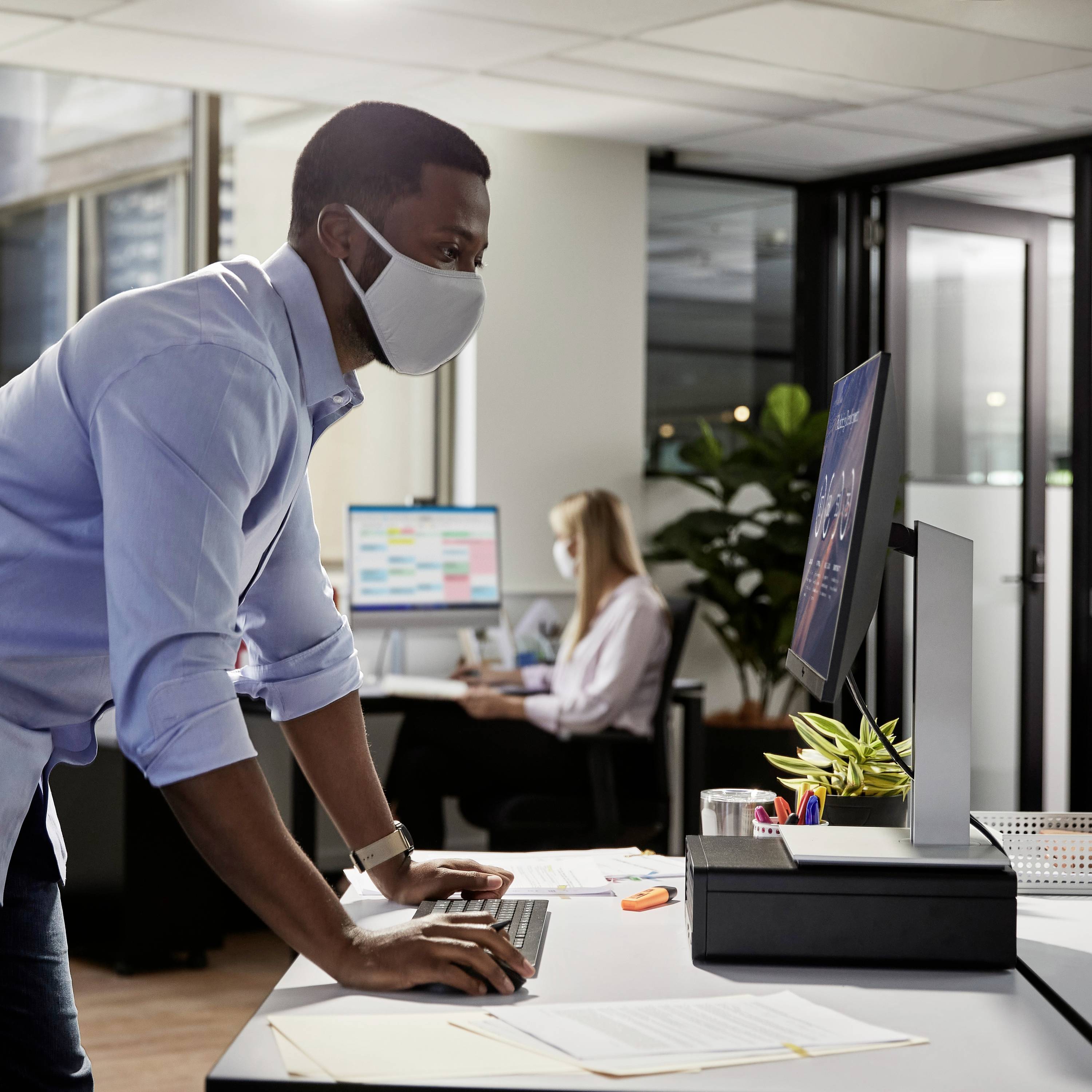 Een man met een gezichtsmasker werkt aan een computer in een modern kantoor. Op de achtergrond zit een vrouw aan een bureau.