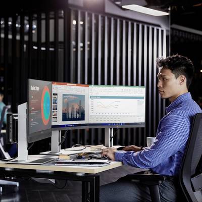 Een man werkt aan twee monitors met diagrammen en tabellen in een modern kantoor. Naast hem liggen documenten en een computer.