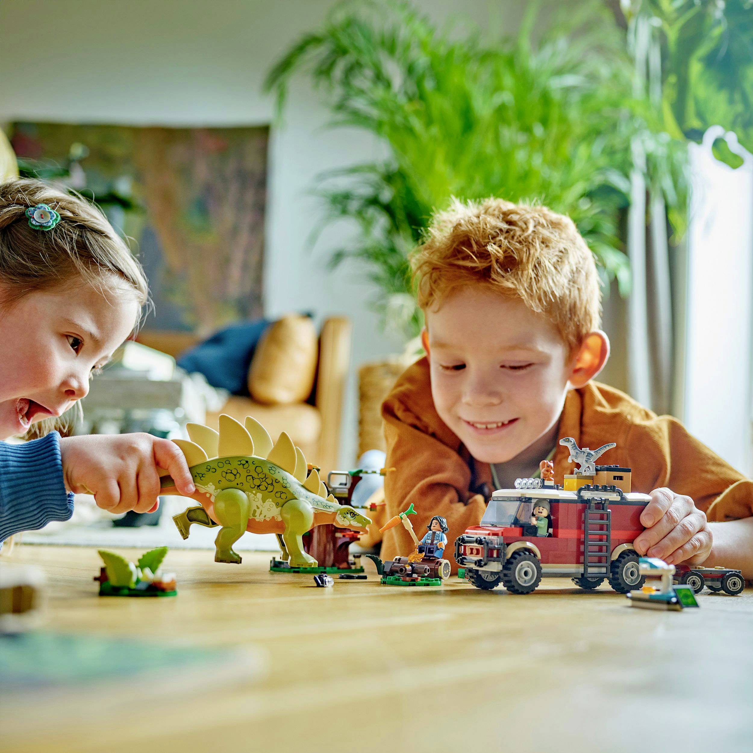 Twee kinderen spelen enthousiast met dinosaurusspelletjes en een speelgoedauto op een tafel. Op de achtergrond zijn planten zichtbaar.