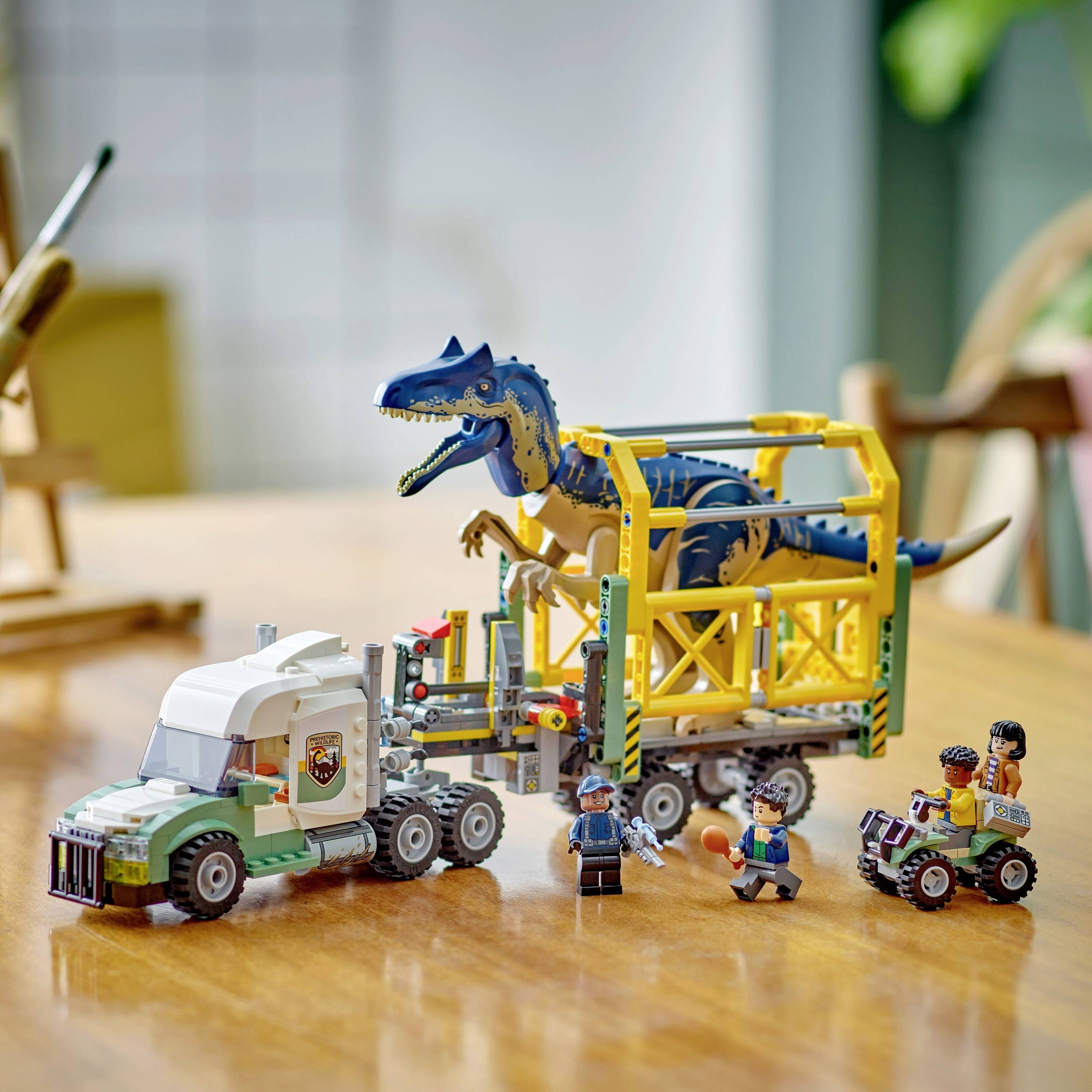 Speelgoedset op een tafel: Een dinosaurus in een transportkooi op een vrachtwagen. Ernaast staan verschillende speelfiguren en een kleine jeep.