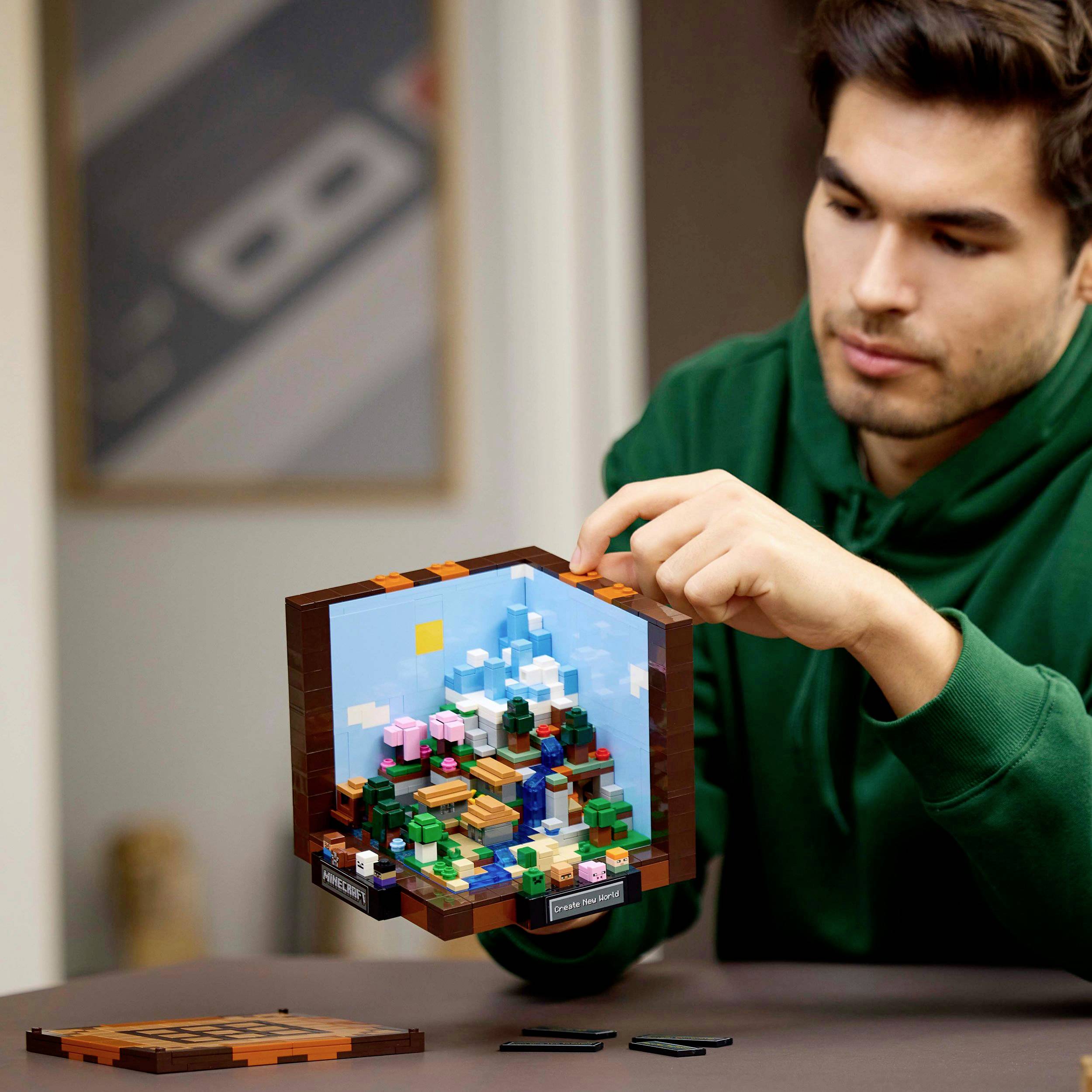 Een man in een groene trui bekijkt een miniatuurmodel met kubussen en landschap, dat gepresenteerd wordt met een open doos.