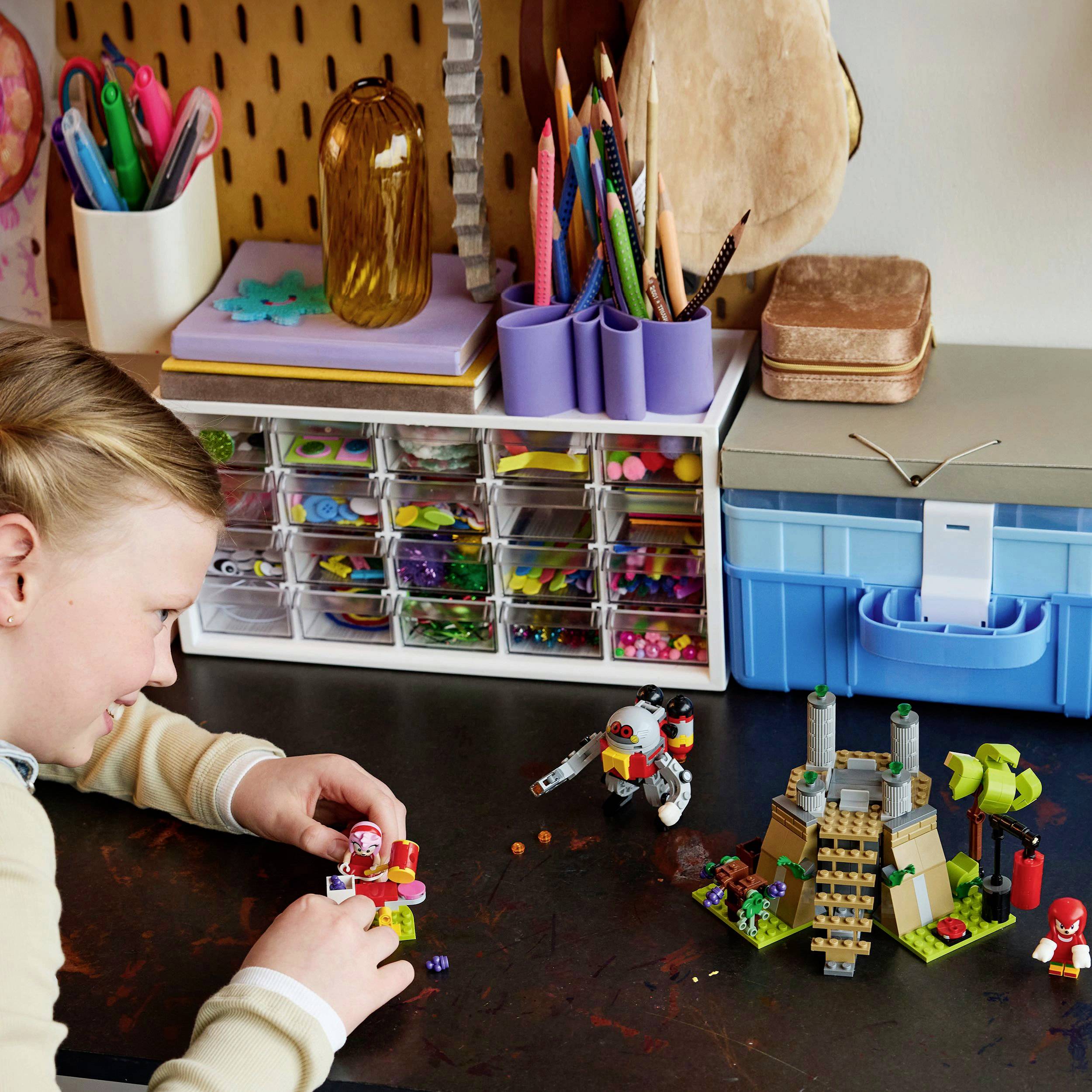 Een kind zit aan een tafel en speelt met LEGO-blokken, omringd door knutselmateriaal. Op de achtergrond zijn dozen en stiften zichtbaar.