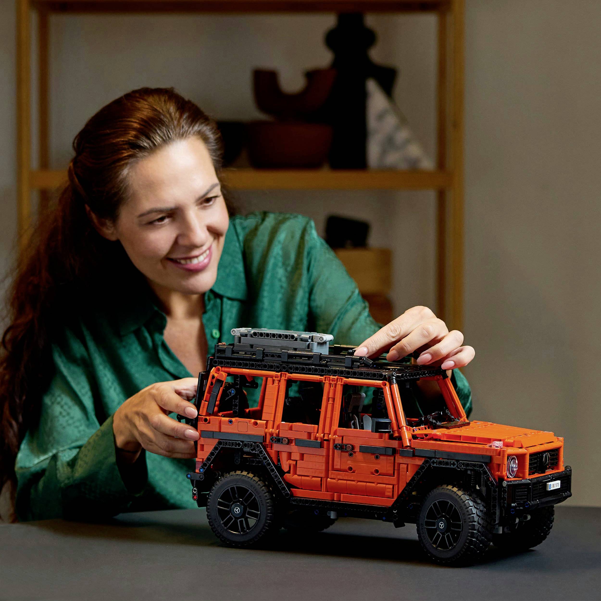 Een vrouw in een groene blouse glimlacht terwijl ze een modelauto in de vorm van een terreinwagen controleert. Op de achtergrond staat een rek.