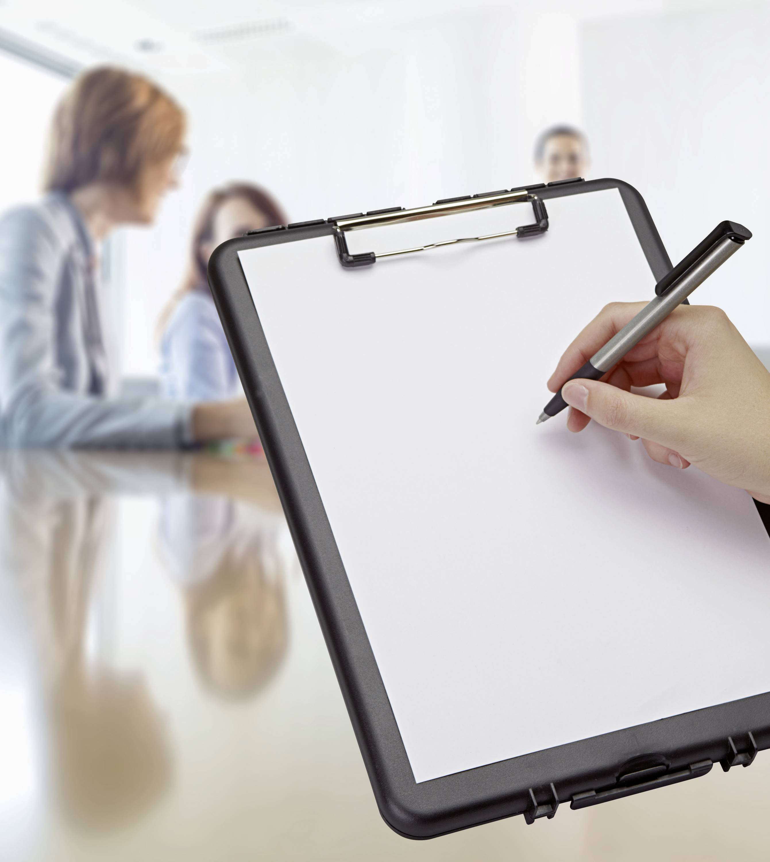 Een hand schrijft op een klembord met wit papier, waarbij drie personen vaag op de achtergrond in een vergadering zitten.