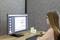 Een vrouw zit aan het bureau en bekijkt een document op een computermonitor. Ze draagt een bril en heeft lang haar.