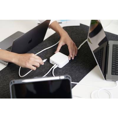 Een persoon steekt een oplaadkabel in een witte USB-hub die op een tafel naast twee laptops en een tablet ligt.