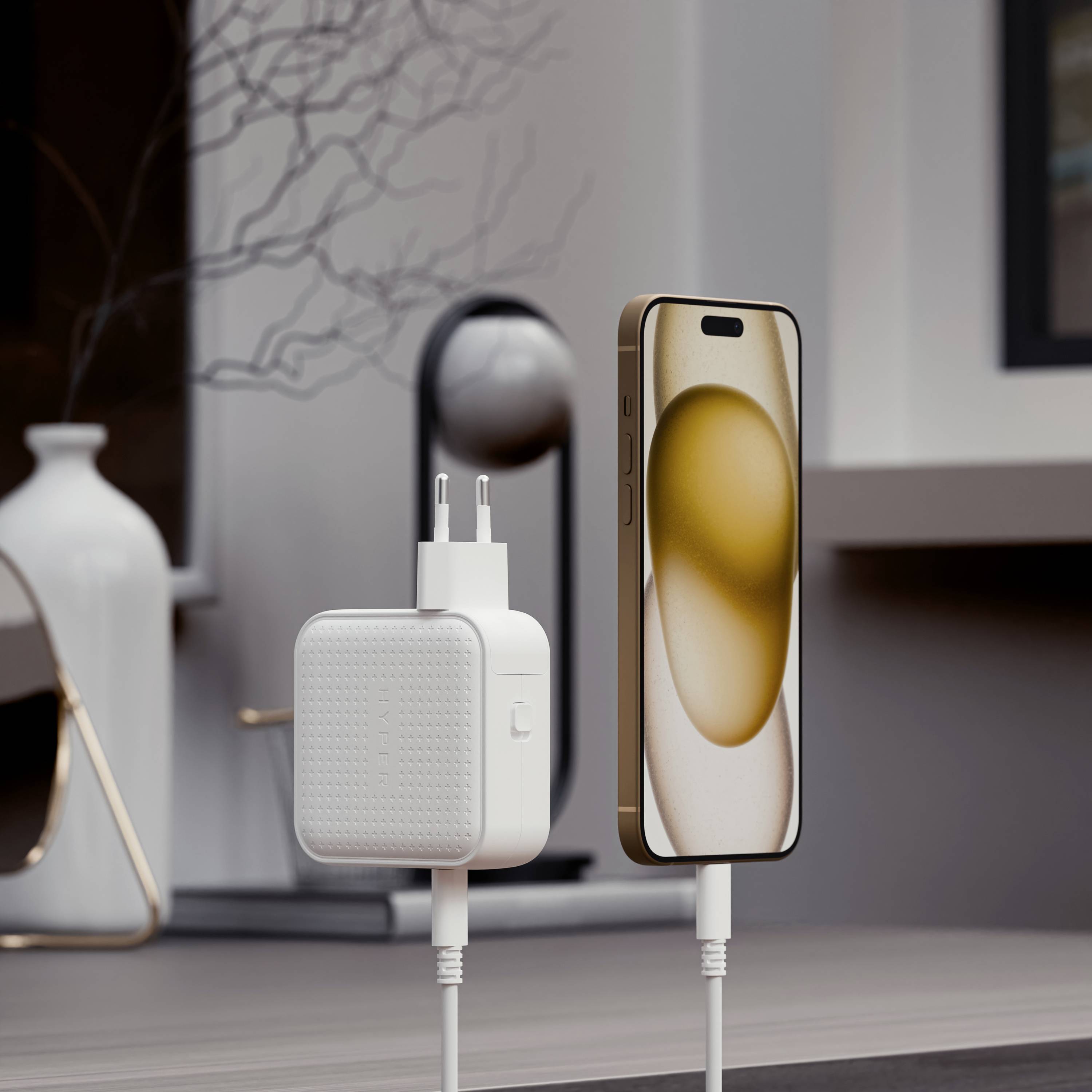 Een smartphone staat rechtop naast een oplader op een tafel, op de achtergrond is een decoratieve vaas zichtbaar.