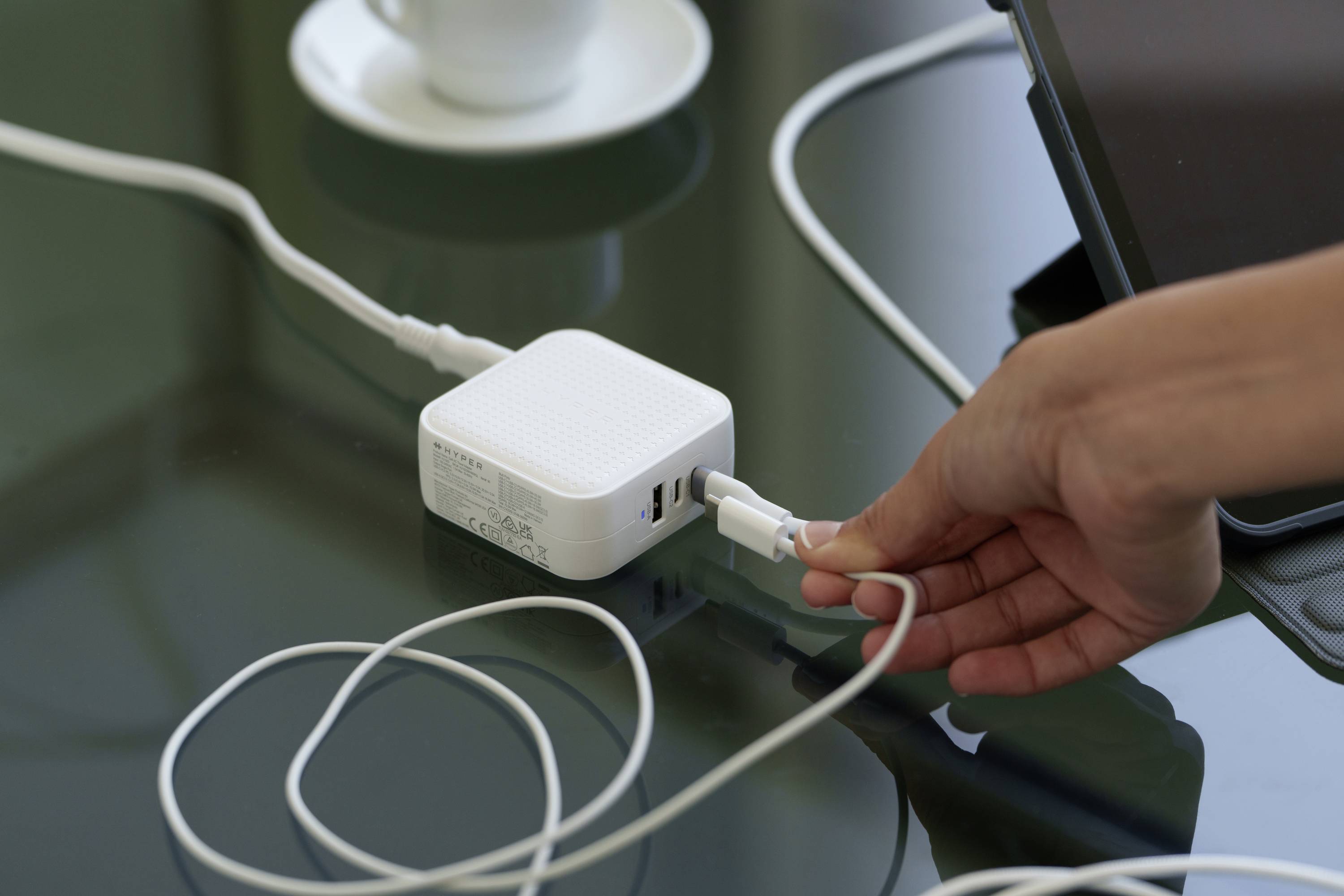 Een hand steekt een kabel in een wit oplaadapparaat op een tafel. Op de achtergrond zijn een kopje en een tablet zichtbaar.