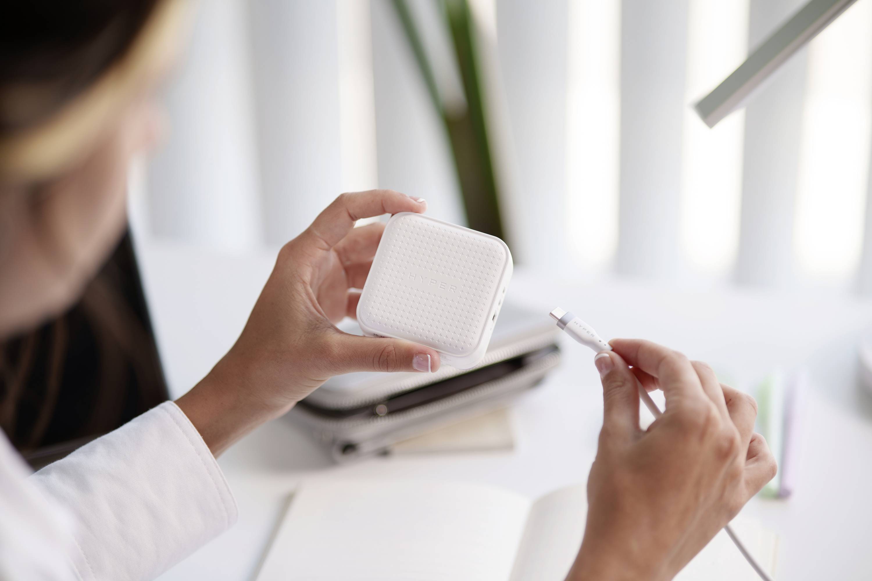 Een persoon houdt een vierkaardig elektronisch apparaat en een oplaadkabel vast, zittend aan een bureau met een notitieboek.