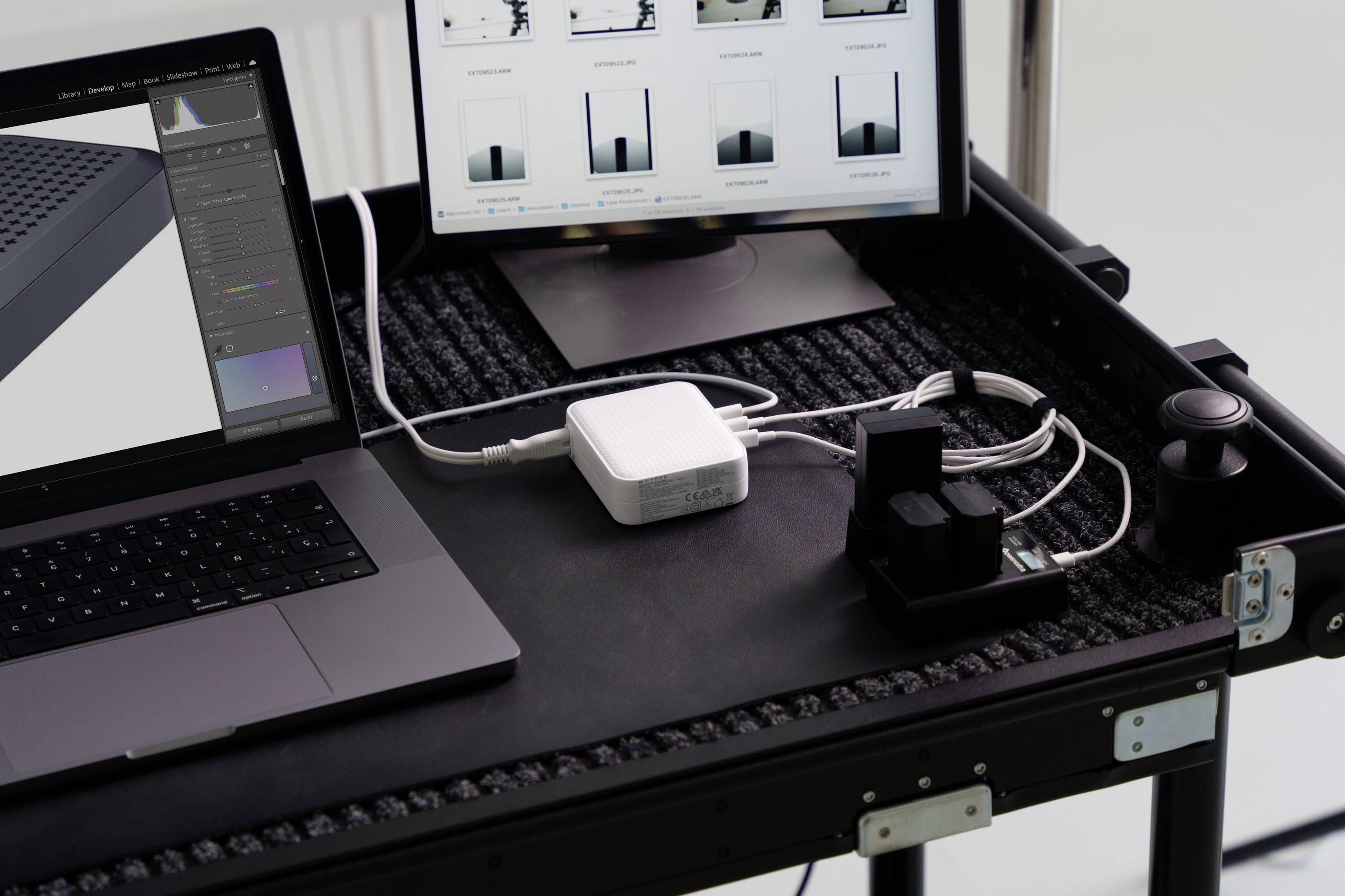 Werkplek met laptop en monitor op een tafel met oplaadstation en kabels. Monitor toont grafieken en fotobewerkingssoftware.