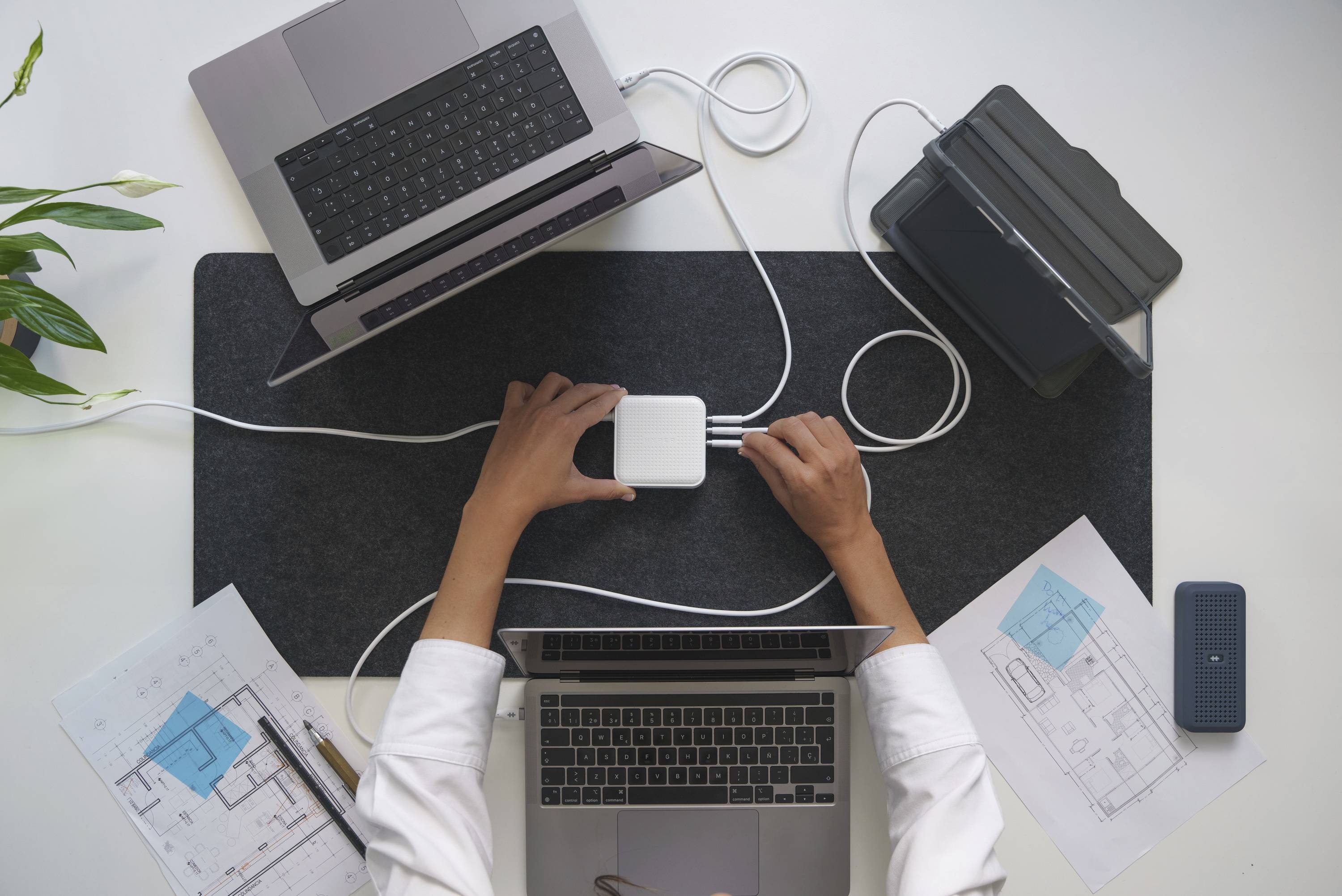 Twee laptops, een tablet en technische tekeningen op een bureau; een persoon verbindt apparaten met een oplader.