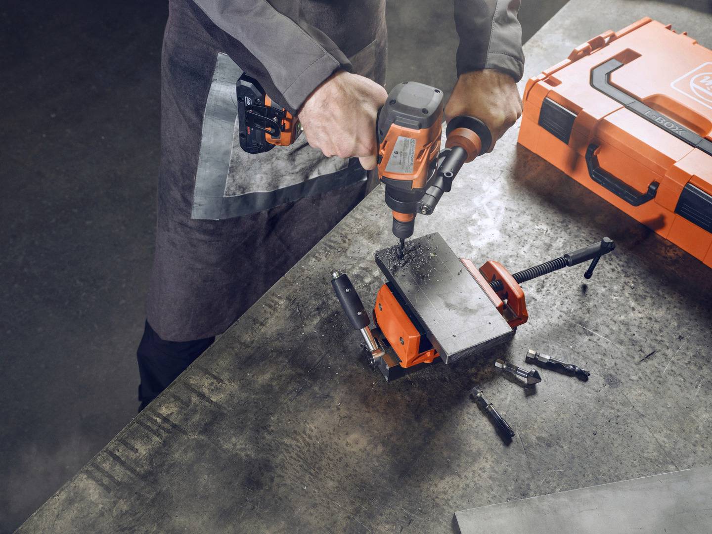 Een persoon boort met een elektrische boormachine in een metalen plaat die in een bankschroef is vastgeklemd. Naast de werkplek staat een oranje gereedschapskoffer.