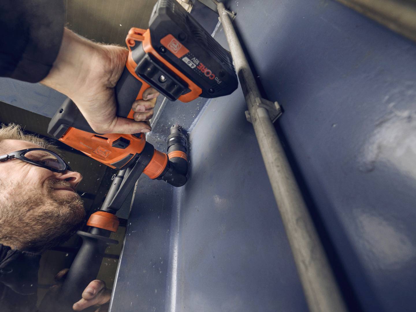 Een man draagt een veiligheidsbril en gebruikt een oranje-zwarte boormachine om in een metalen oppervlak te boren.