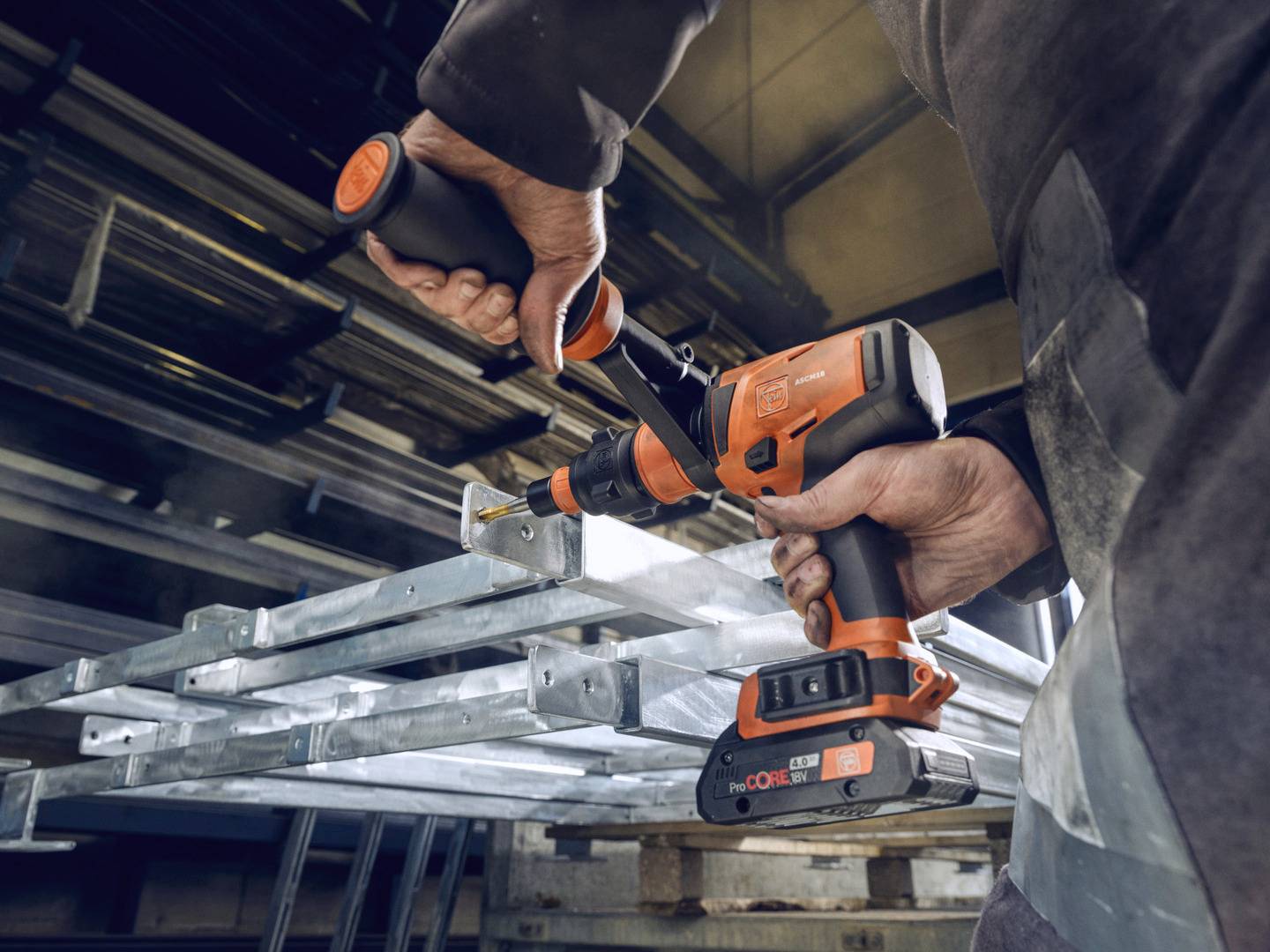 Een persoon gebruikt een oranje-zwarte boormachine om een metalen rek te monteren of te bewerken in een industriële omgeving.