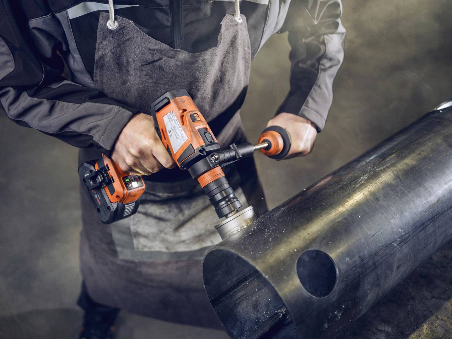 Een persoon in werkkleding gebruikt een oranje boormachine om een groot metalen pijp te bewerken.