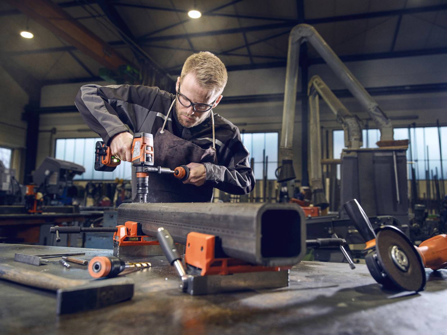 Een man in beschermende kleding bewerkt in een werkplaats een groot metalen pijp met een boormachine. Op de achtergrond zijn gereedschappen te zien.