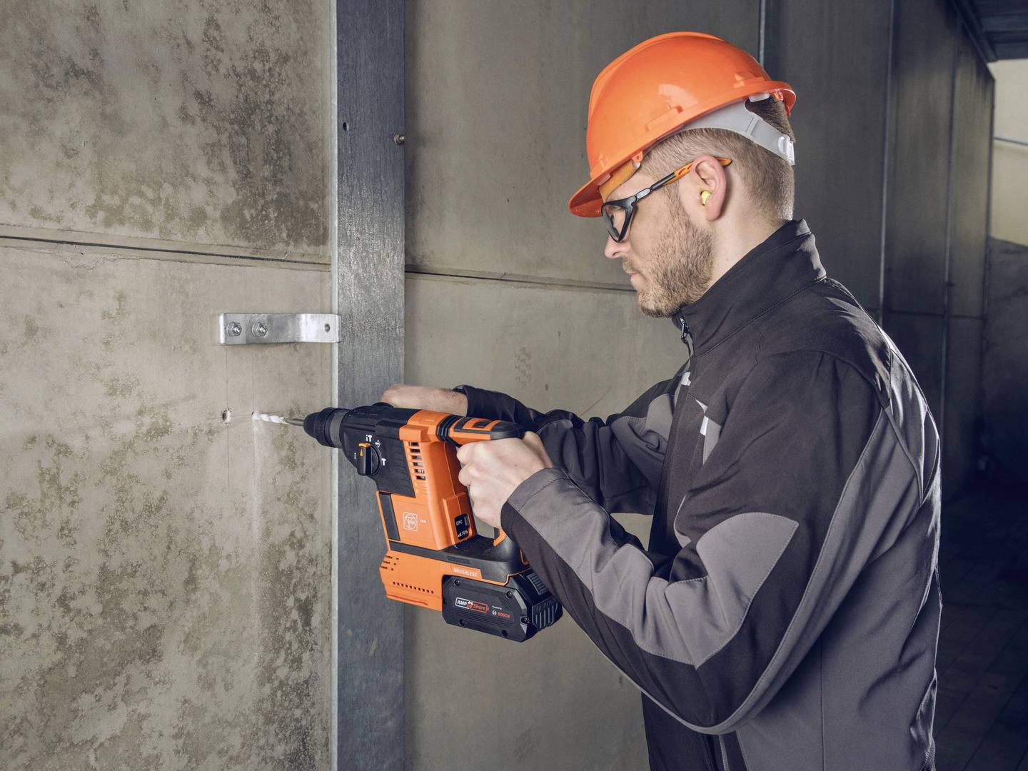 Een arbeider met veiligheidsbril en helm boort met een oranje boorhamer in een betonnen muur.