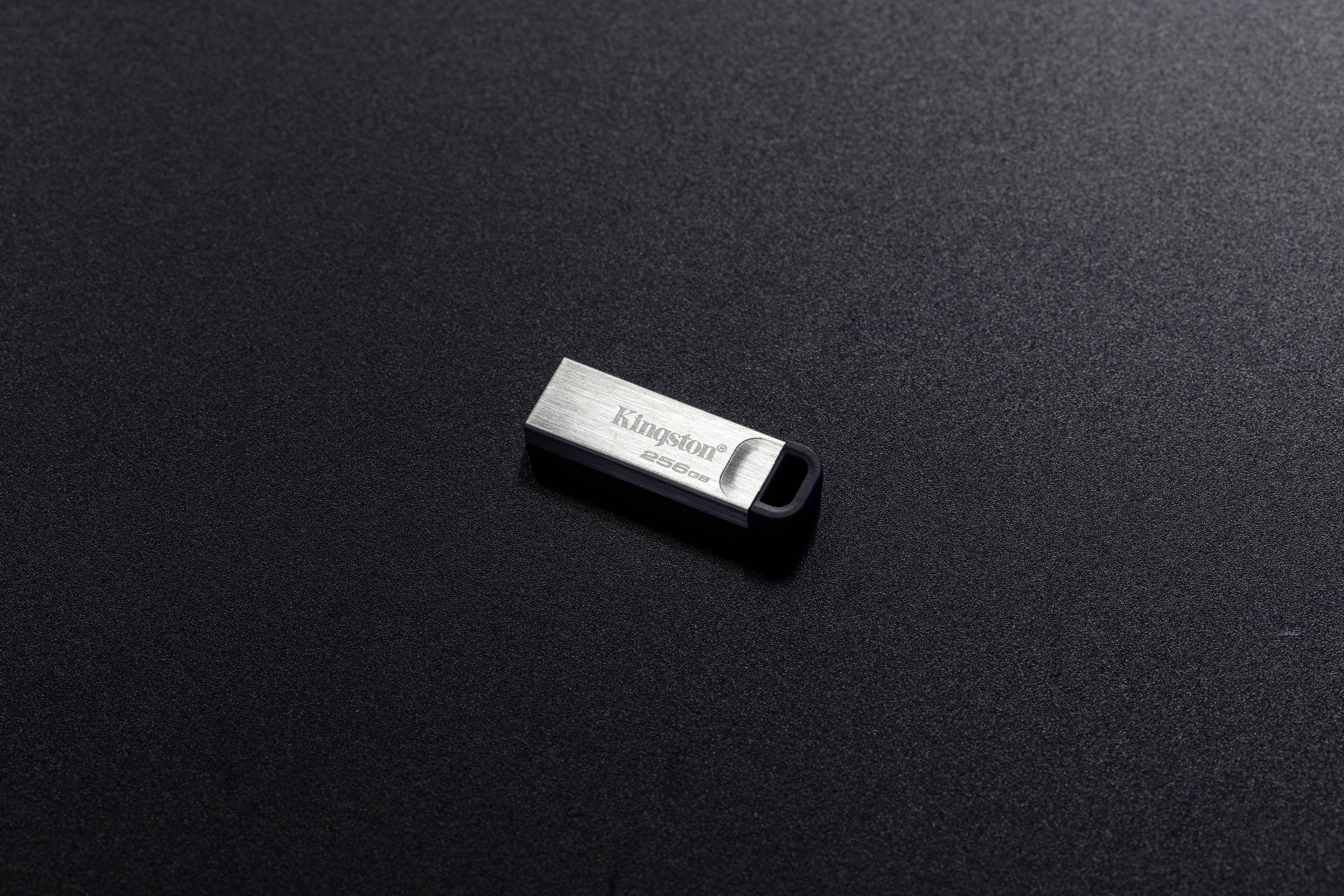 Een kleine USB-stick ligt op een donkere achtergrond. Hij is zilverkleurig en heeft een rechthoekige vorm.