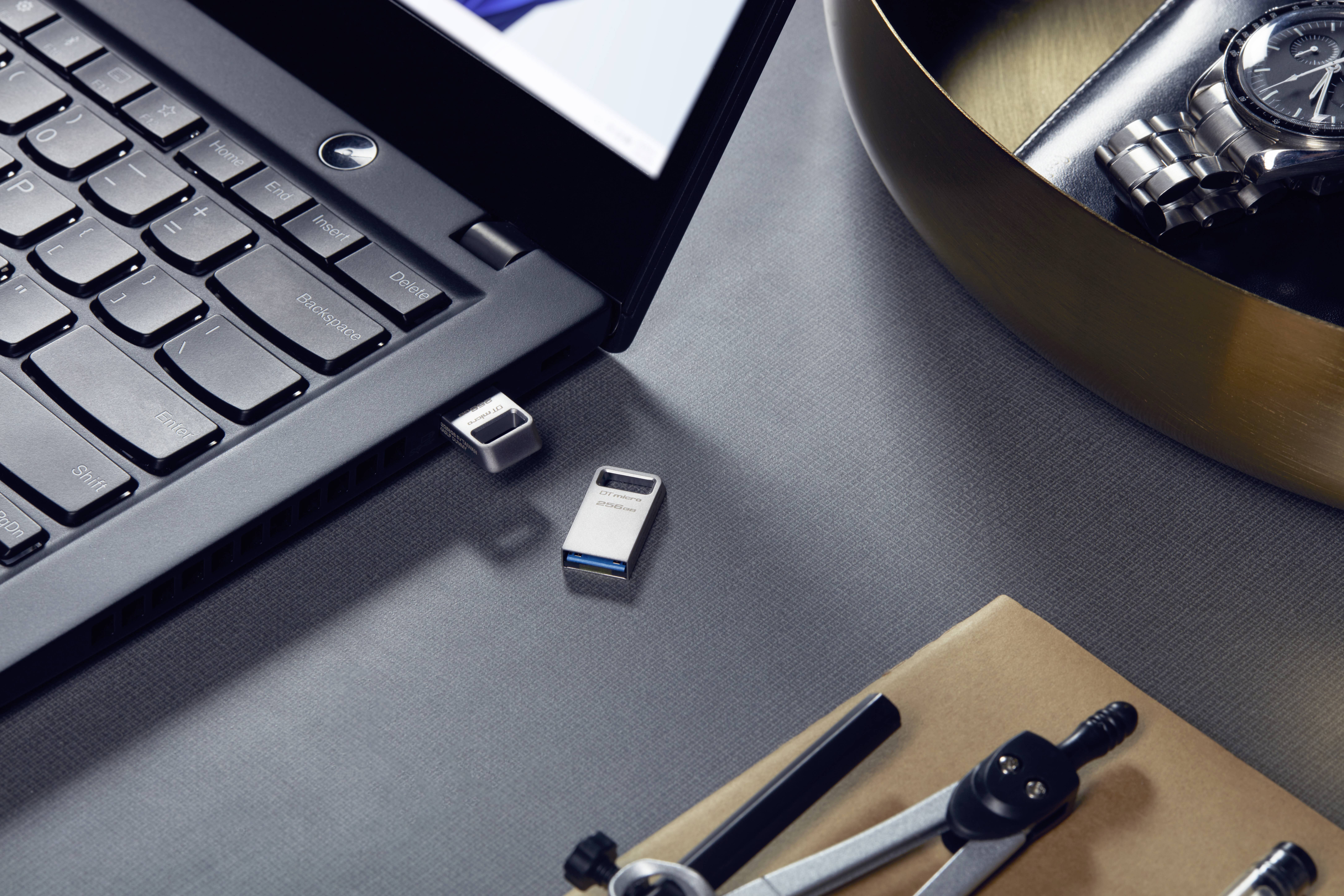 Laptop en USB-stick op bureau met passer en klok op de achtergrond.