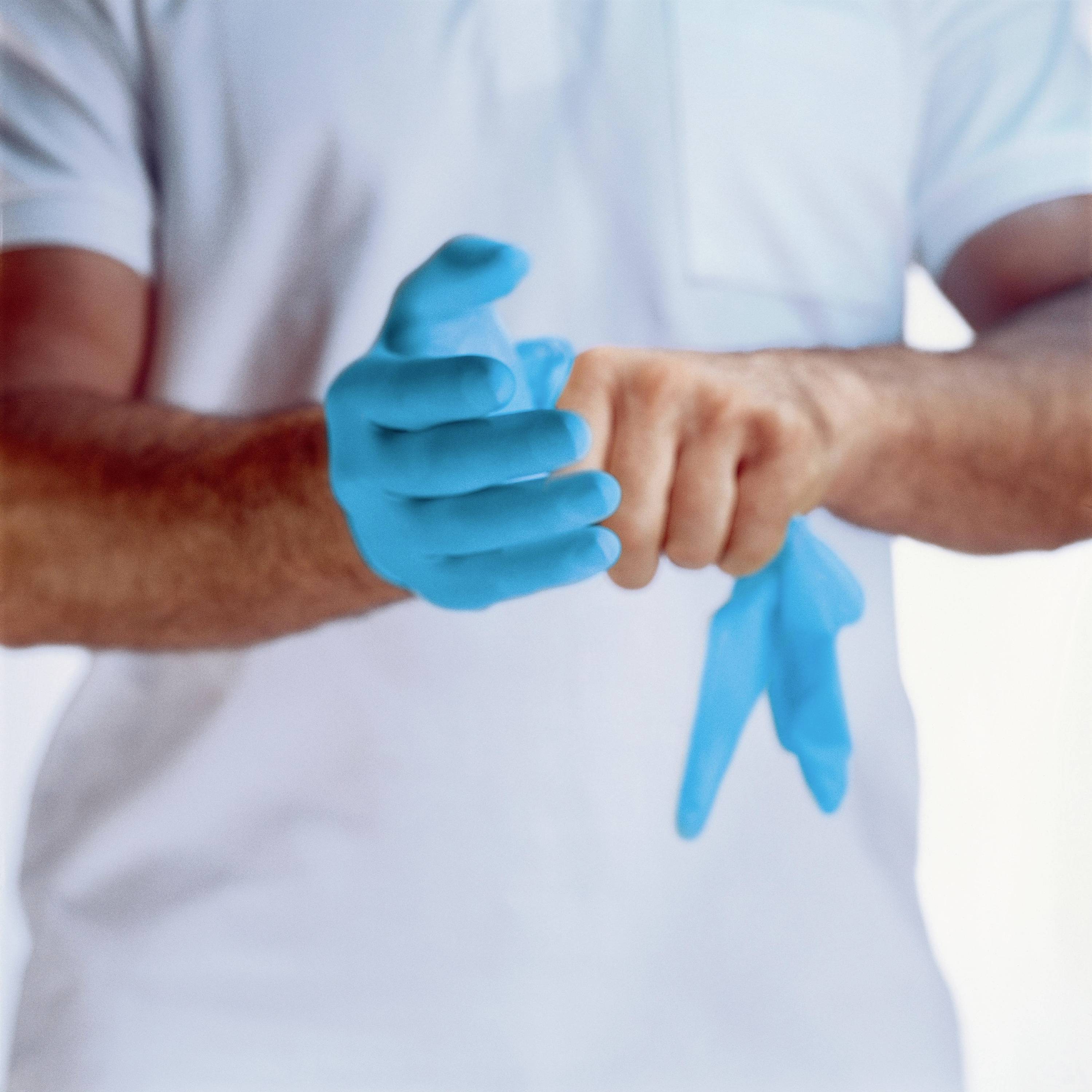 Een persoon trekt blauwe wegwerphandschoenen aan. Hij draagt een wit T-shirt.